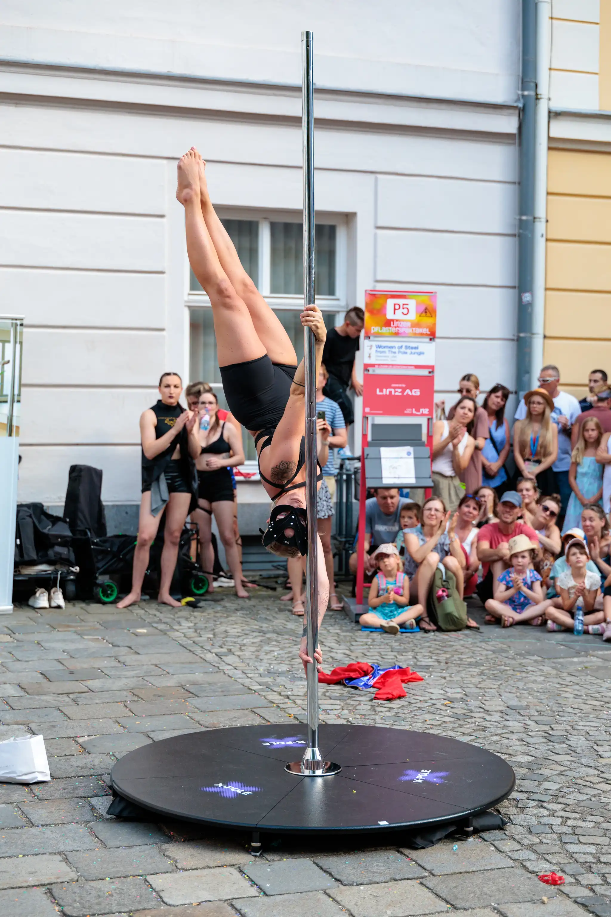 Frau in schwarzer Sportkleidung bei einer Pole-Dance-Performance im Freien, kopfüber an einer tragbaren Stange mit Publikum