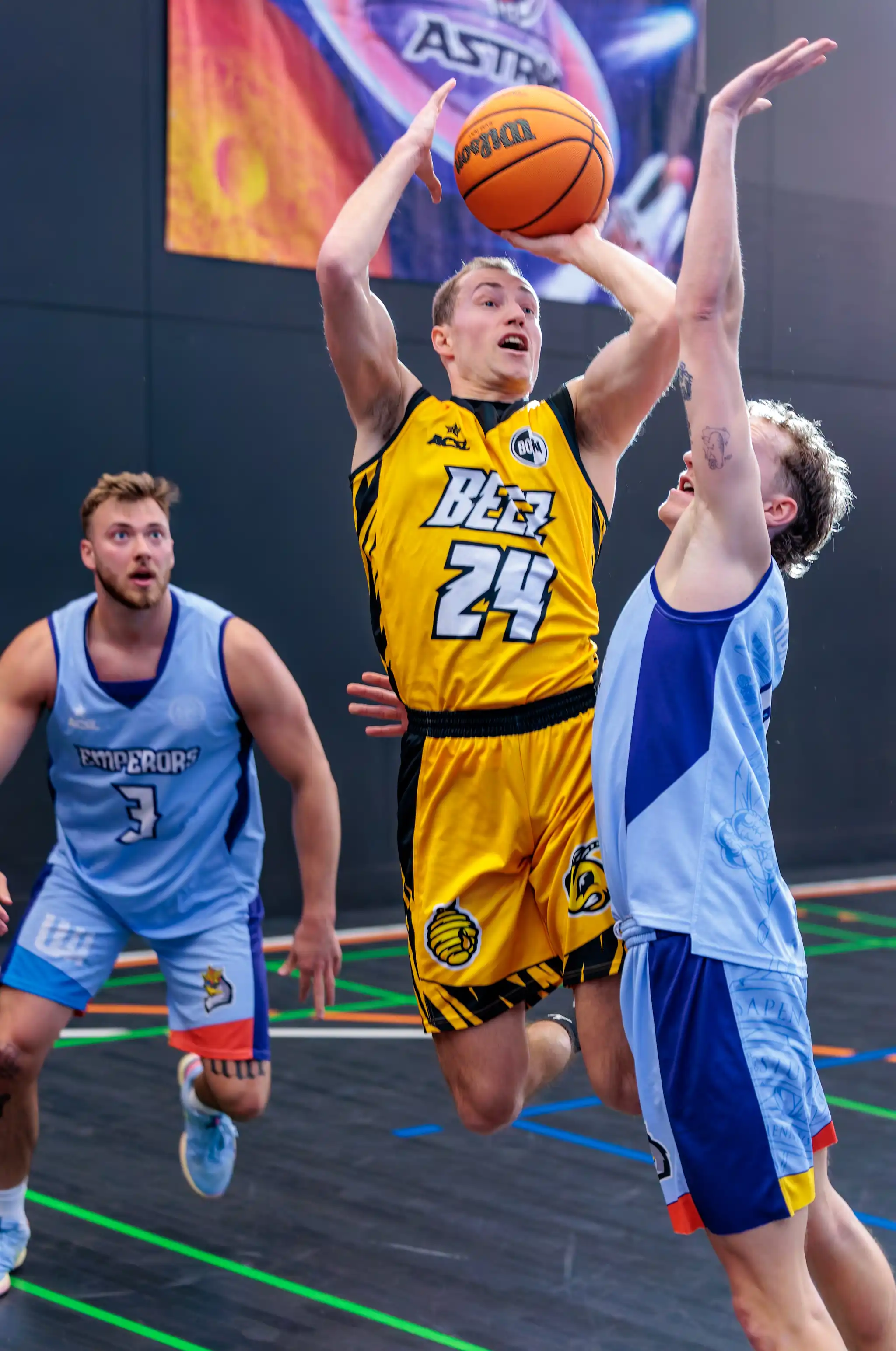 Ein Basketballspieler in gelbem Trikot springt beim Wurf auf den Korb, von einem Gegenspieler verteidigt 