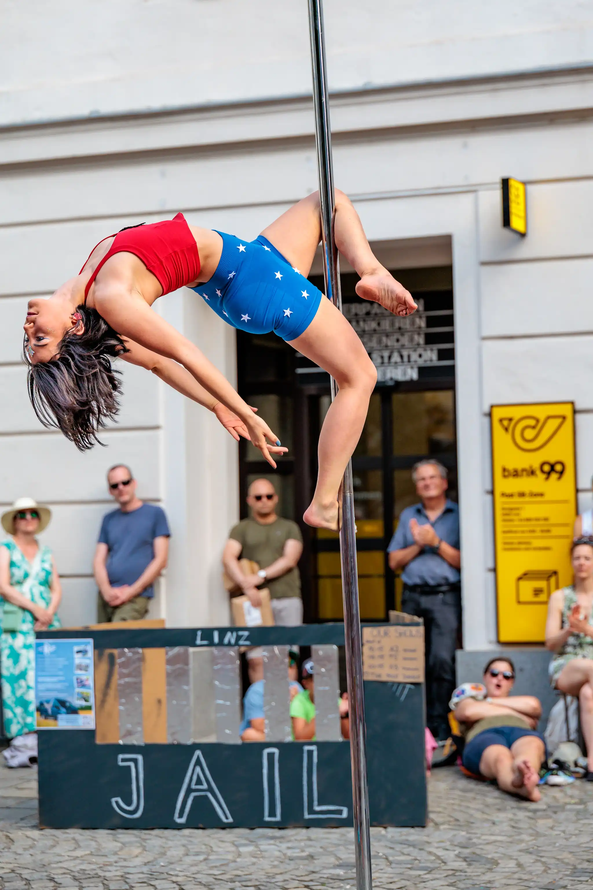 Junge Frau in einem rot-blauen Wonder Woman Kostüm führt im Freien eine akrobatische Pole-Dance-Figur auf, umgeben von Zuschauern und einem Requisit mit der Aufschrift "LINZ JAIL"