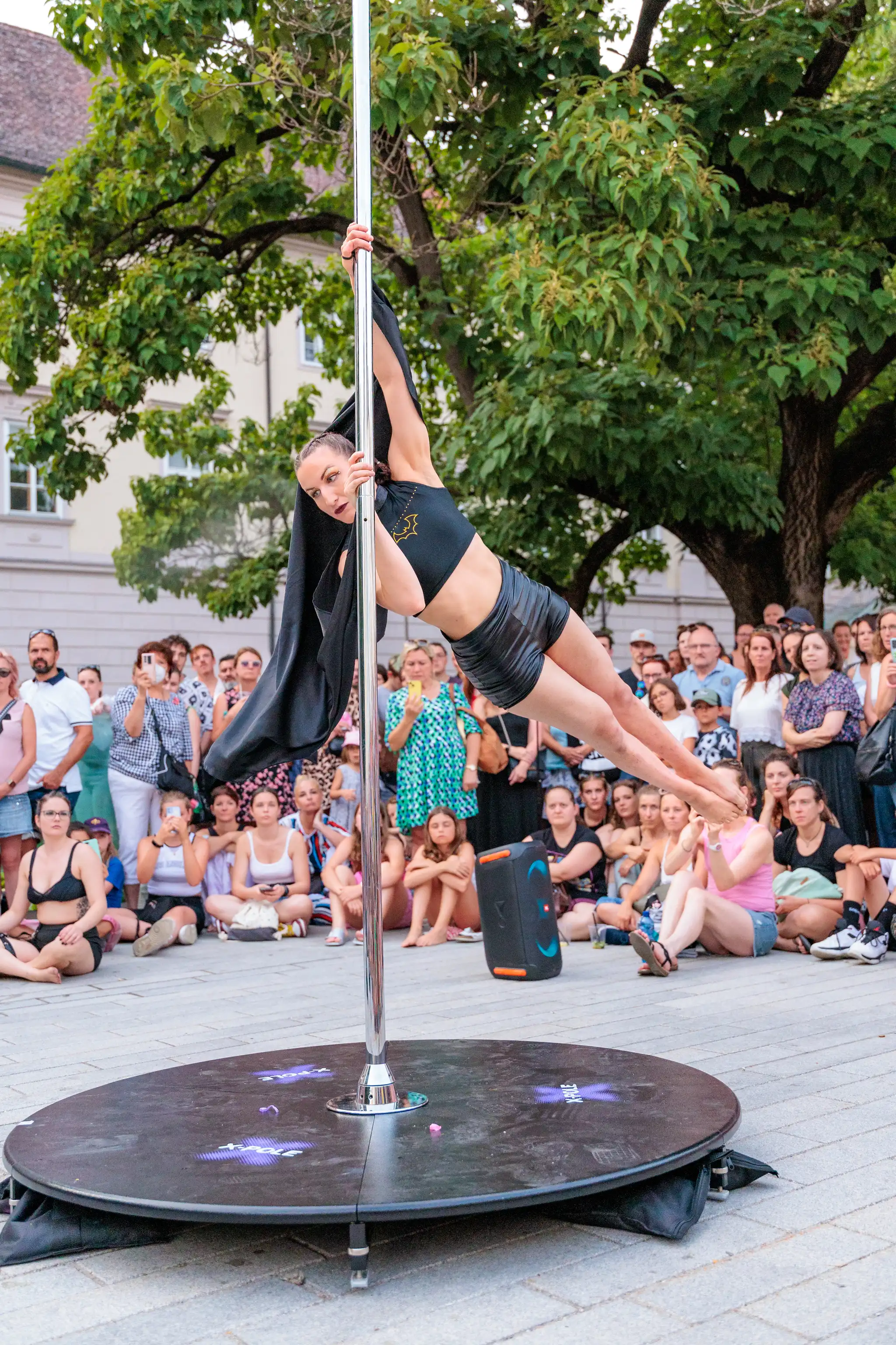 Outdoor Poledance Show während des Pflasterspektakels in Linz: Eine Frau in schwarzem Sportoutfit mit Fledermaus-Logo zeigt eine horizontale Akrobatik-Figur vor einem Publikum