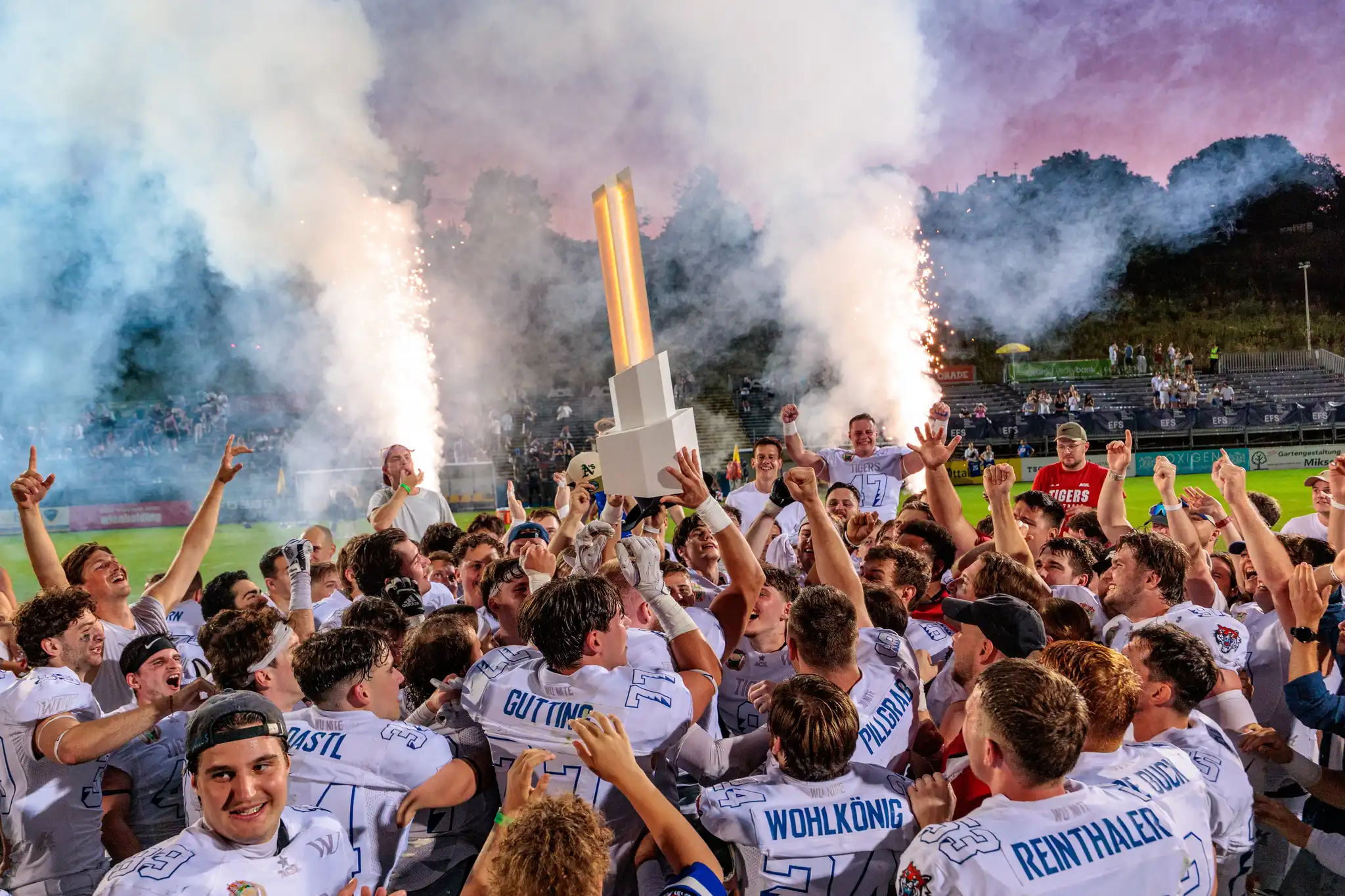 Amerikanische Footballspieler in weißen Trikots feiern ausgelassen ihren Meisterschaftssieg mit einem leuchtenden Pokal und Pyrotechnik in einem Stadion 