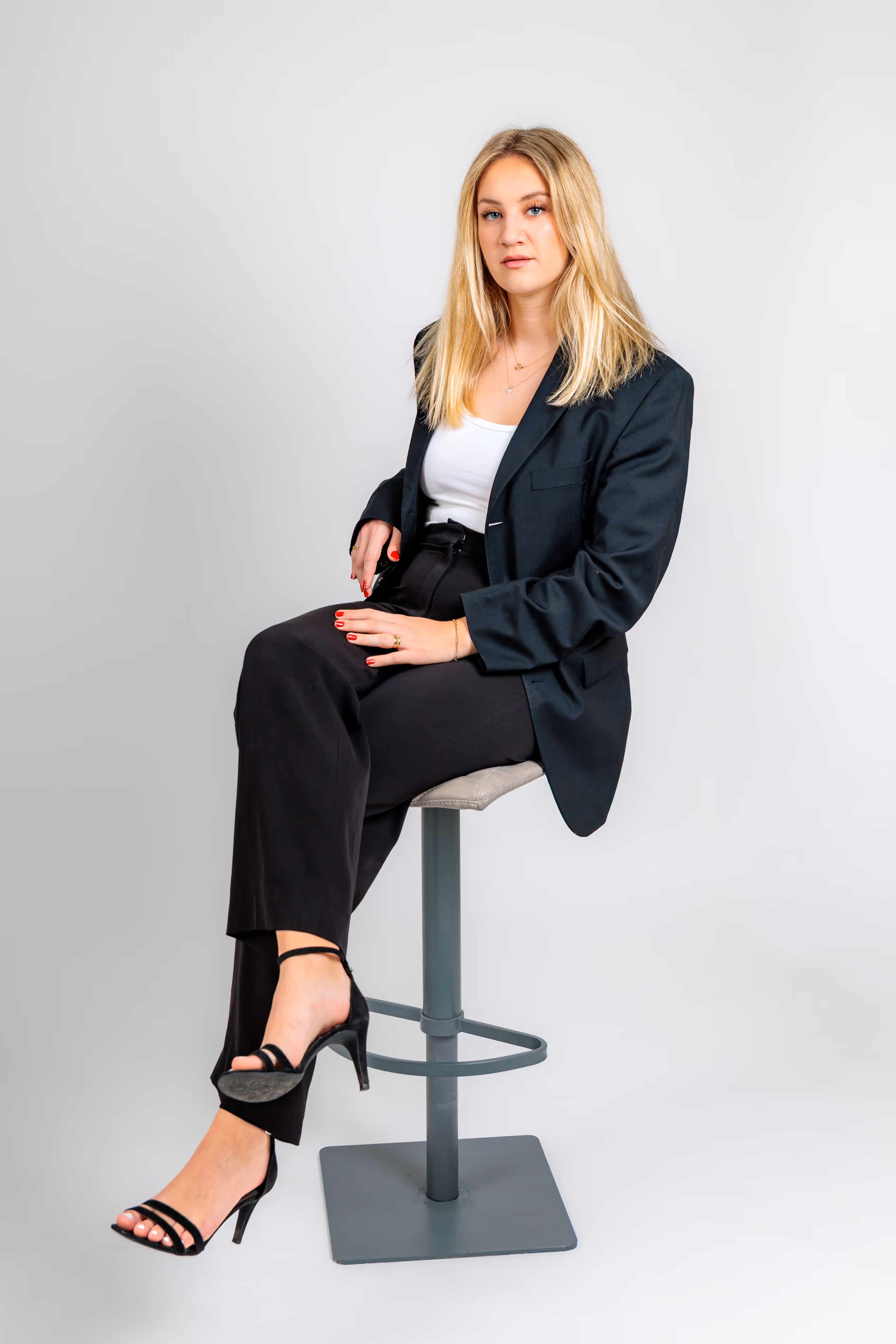 Eine junge blonde Frau im schwarzen Hosenanzug sitzt für ein Business-Fotoshooting selbstbewusst auf einem Barhocker vor einem weißen Hintergrund