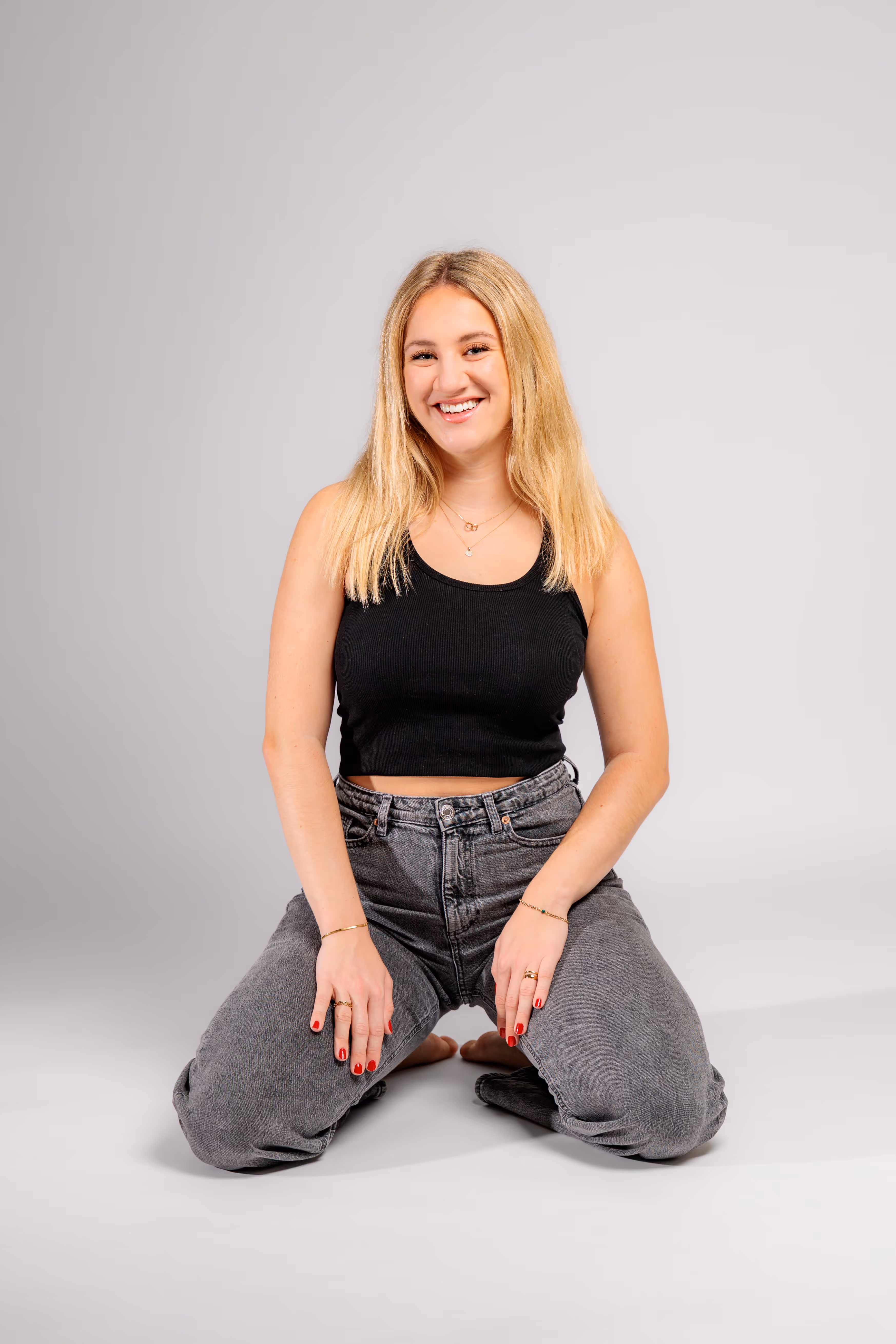 Das Model Anna kniet in einem lässigen Outfit mit Jeans und schwarzem Top auf dem weißen Boden des Fotostudios und lächelt in die Kamera