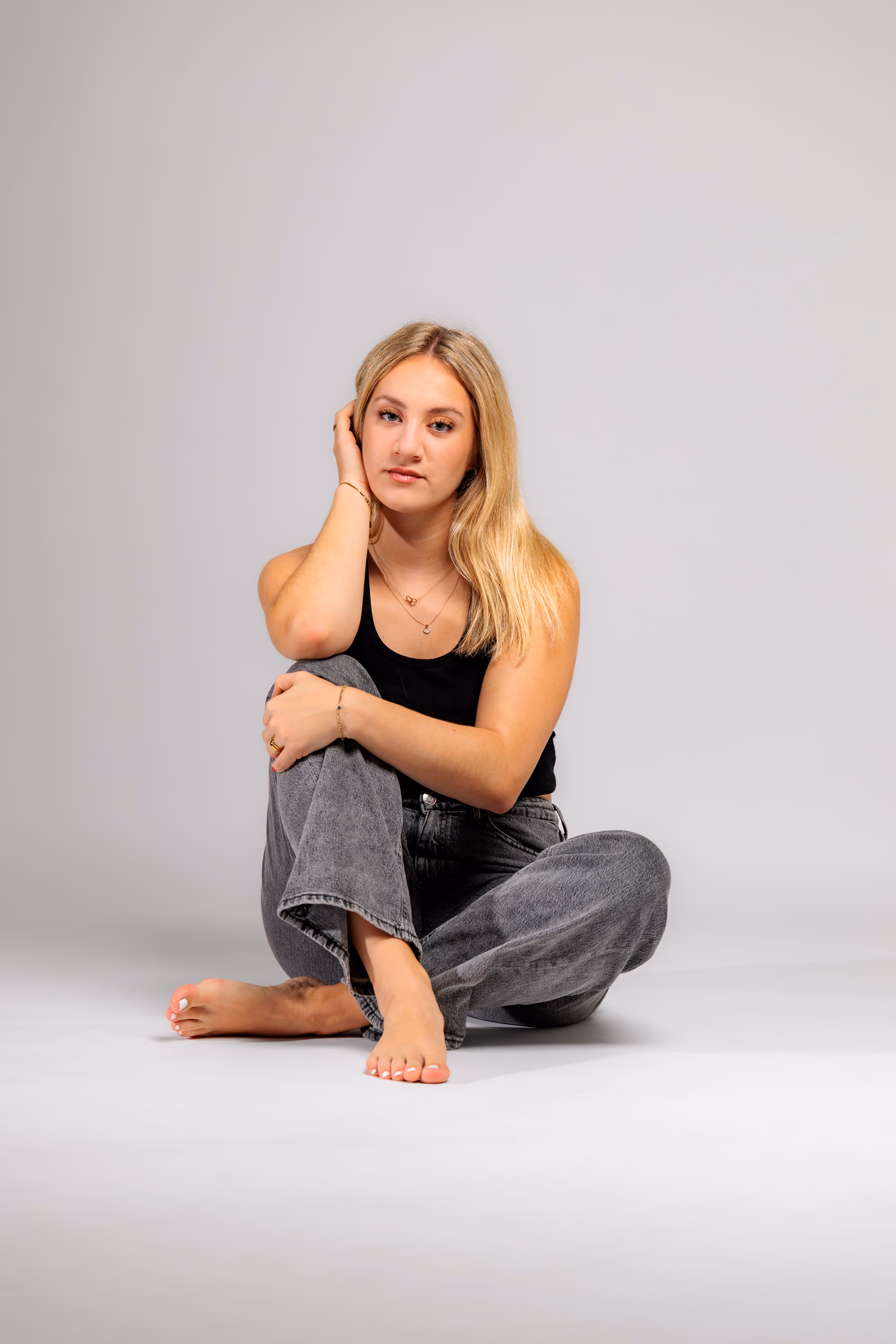 Ganzkörperaufnahme von Model Anna, die in einem lässigen Outfit mit grauer Jeans und schwarzem Top barfuß auf dem Boden des Fotostudios sitzt und in die Kamera blickt