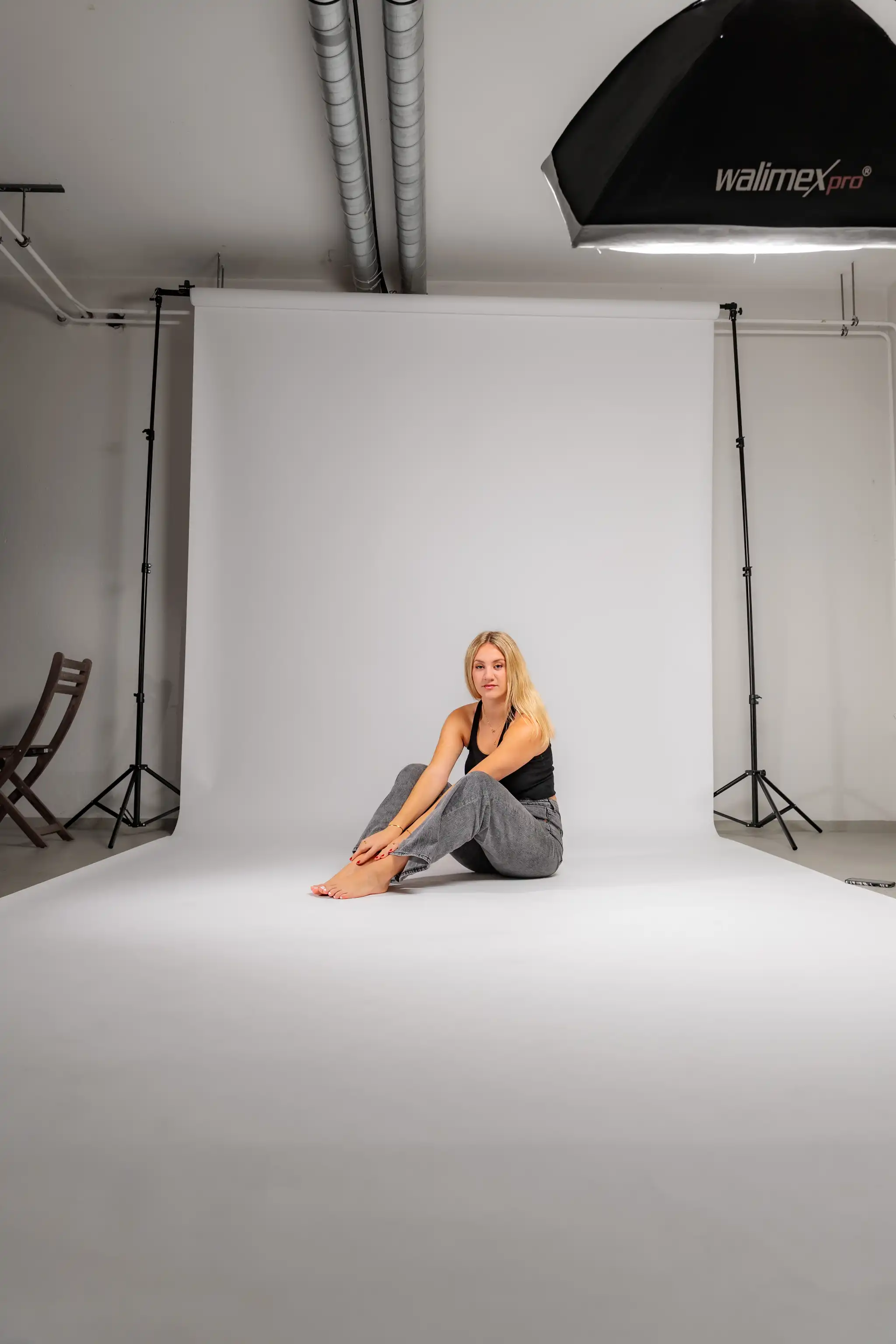 Ein Blick hinter die Kulissen eines Studio-Fotoshootings zeigt Model Anna, die barfuß auf einer weißen Hohlkehle sitzt, umgeben von Stativen und Beleuchtungstechnik