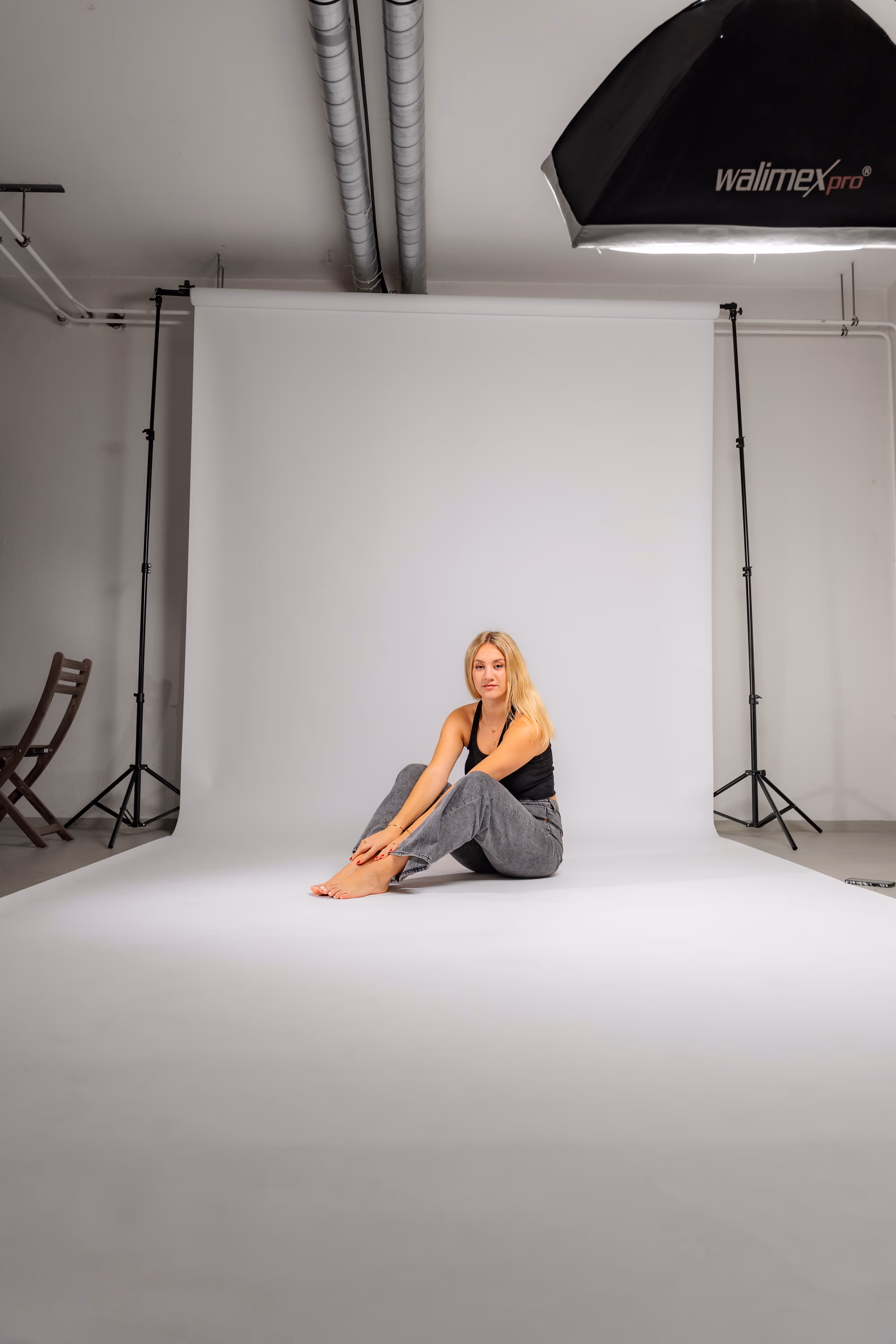 Ein Blick hinter die Kulissen eines Studio-Fotoshootings zeigt Model Anna, die barfuß auf einer weißen Hohlkehle sitzt, umgeben von Stativen und Beleuchtungstechnik