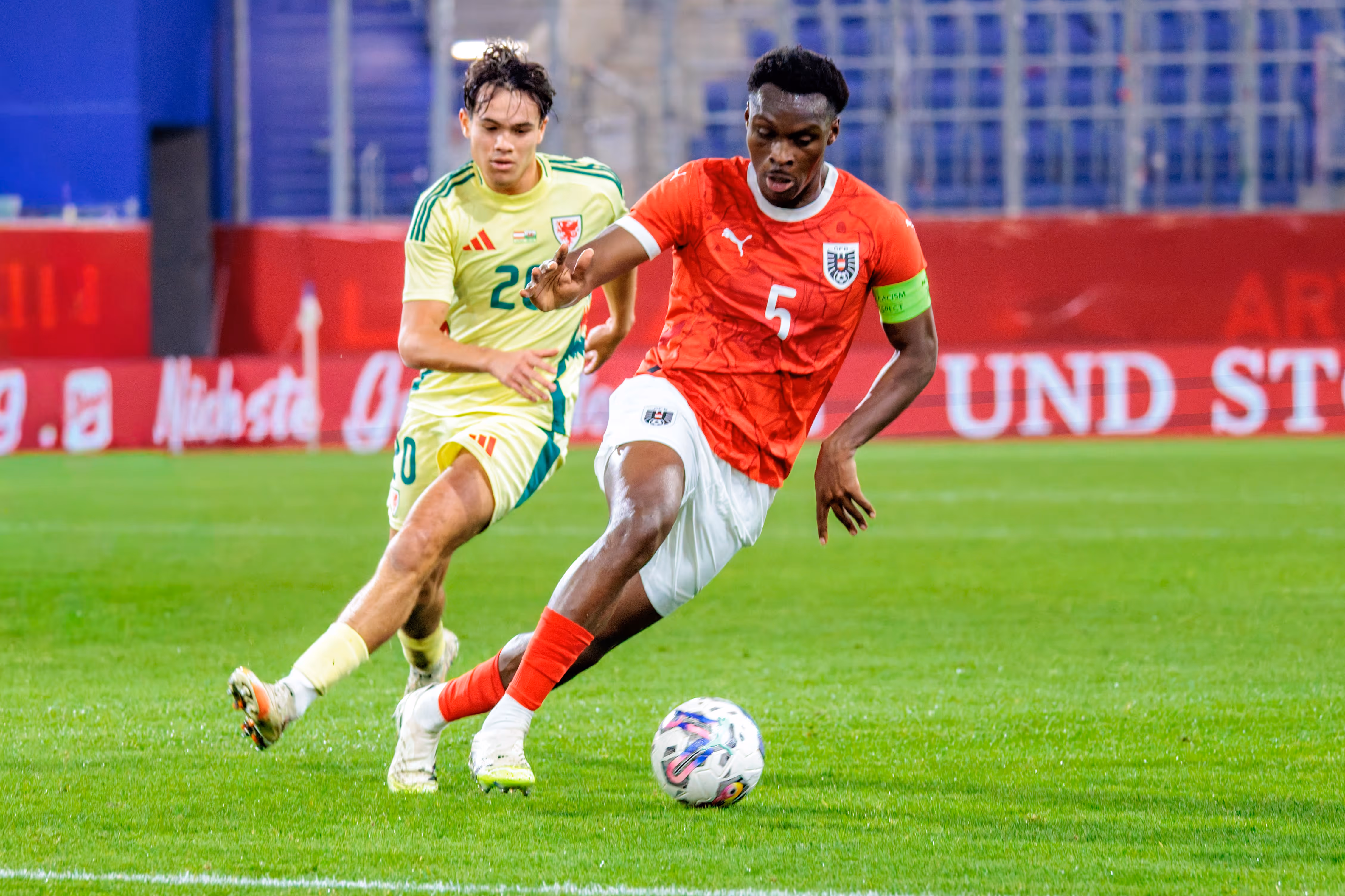 Samson Baidoo (Nummer 5, Österreich) im roten Trikot führt den Ball im Zweikampf mit Gabriele Biancheri (Nummer 20, Wales) im gelben Trikot