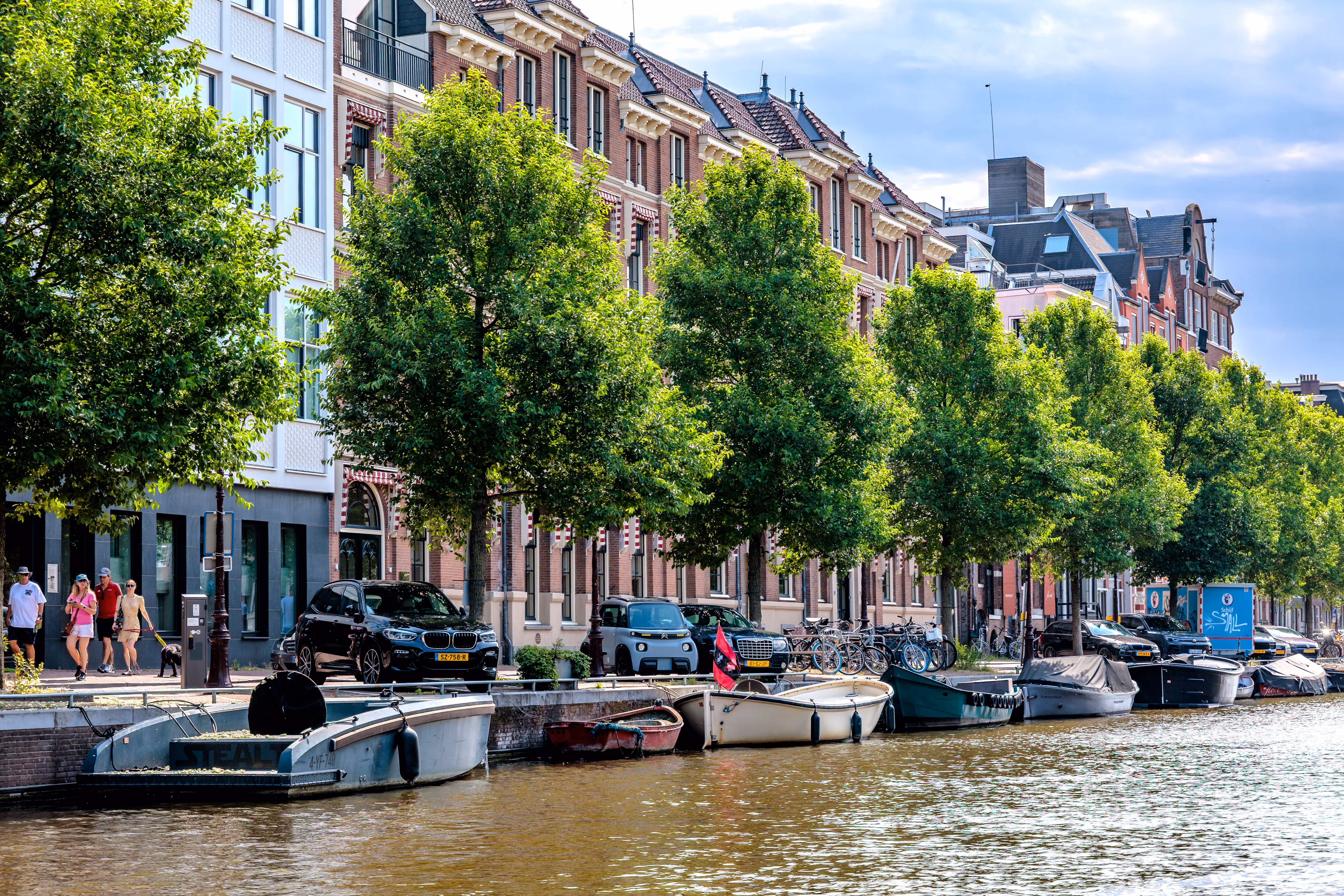 Ein Baumalleen-Kanal in Amsterdam mit historischen Ziegelgebäuden und geparkten Booten bei sonnigem Wetter 