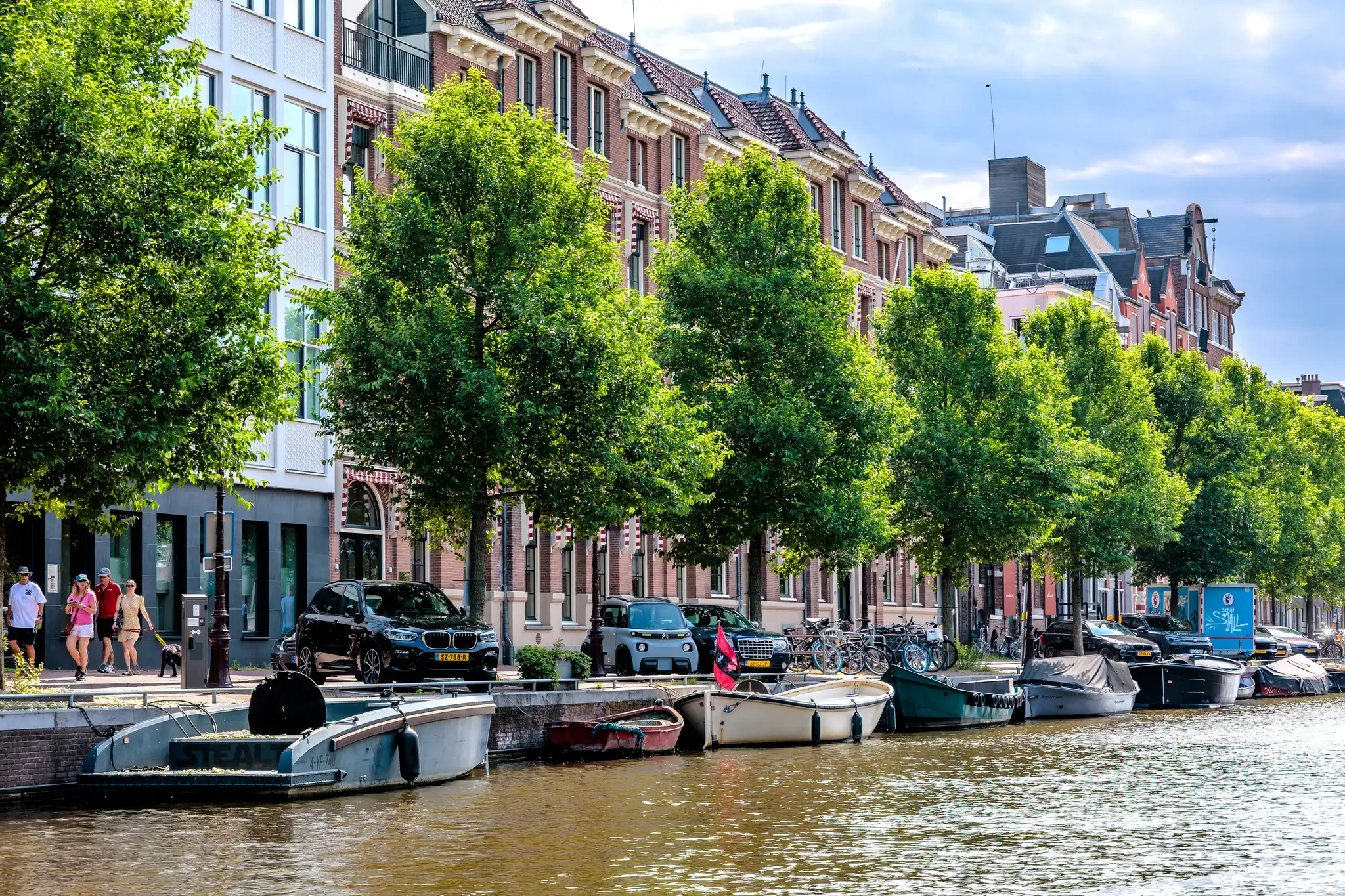 Ein Baumalleen-Kanal in Amsterdam mit historischen Ziegelgebäuden und geparkten Booten bei sonnigem Wetter