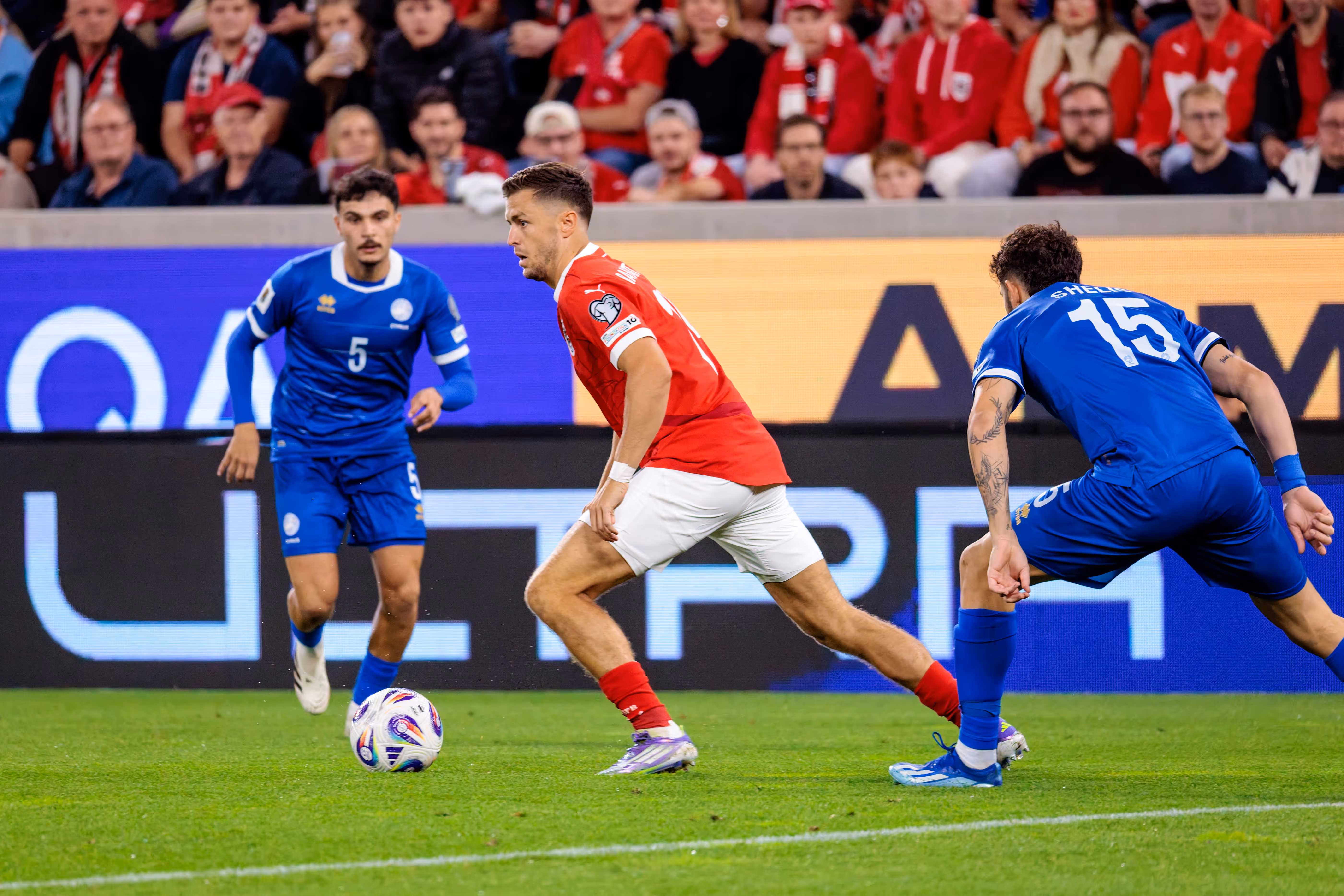 Fußballspieler Christoph Baumgartner im roten Trikot dribbelt den Ball im Zweikampf mit zwei zyprischen Spielern in blauen Trikots während eines Länderspiels