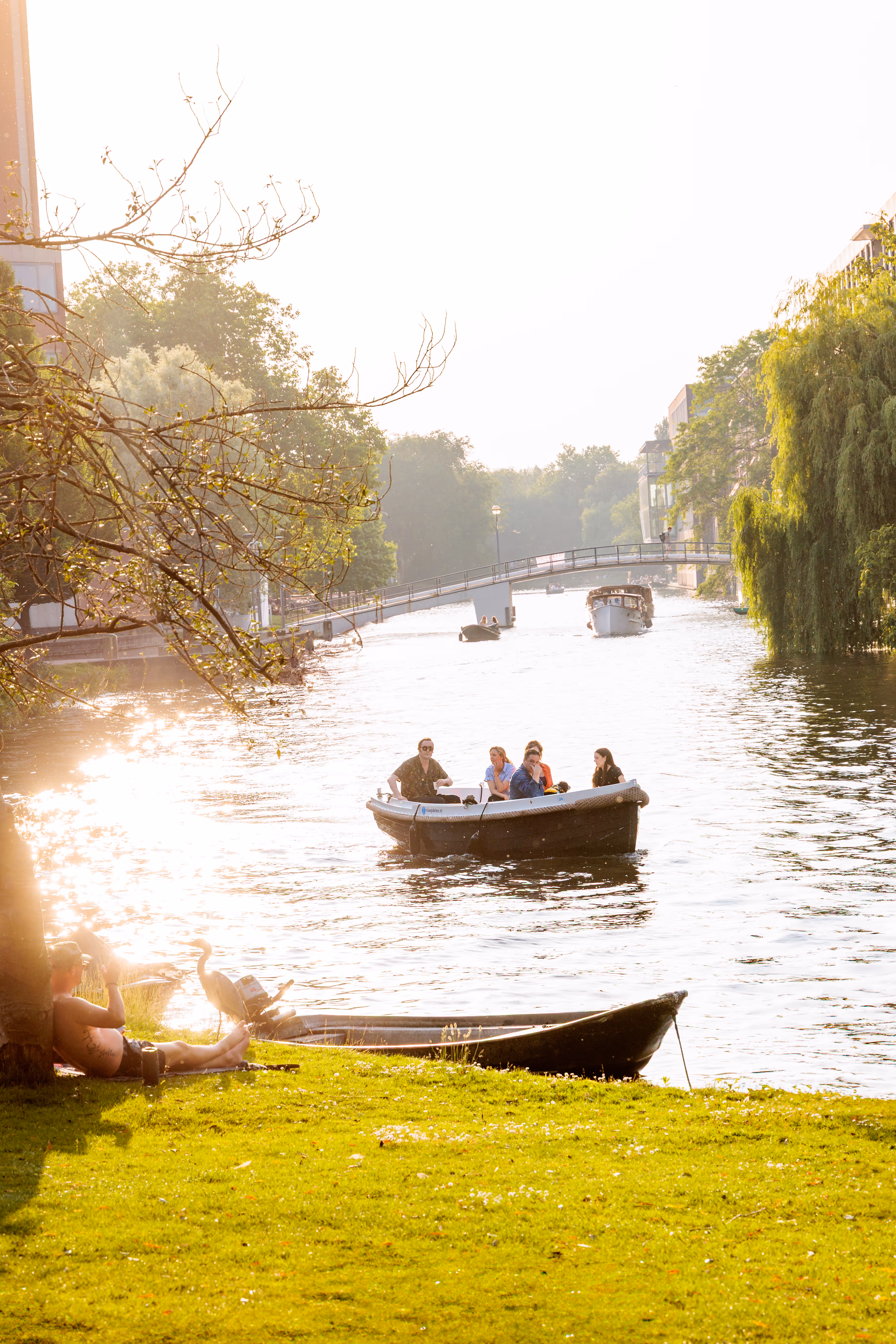 Ein Boot mit Personen gleitet auf einem sonnenbeschienenen Kanal, während am Ufer ein Mann entspannt und ein Reiher danebensteht 