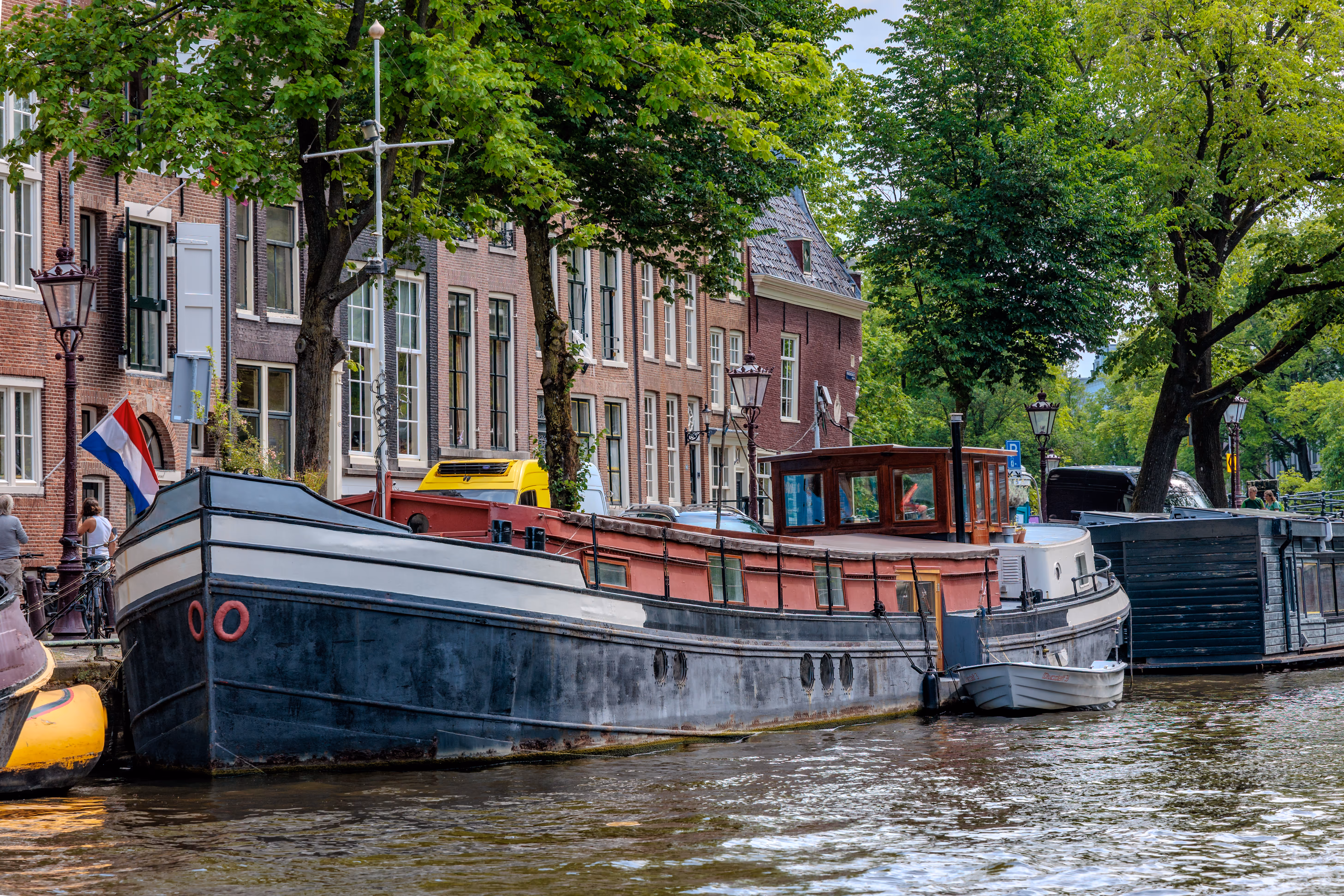 Ein malerisches Amsterdamer Grachtenmotiv mit einem Hausboot vor klassischen Ziegelhäusern und sattgrünen Bäumen 