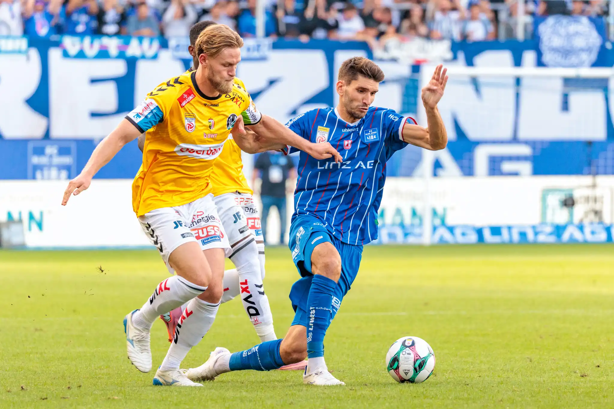 Ein Spieler des SV Ried im gelben Trikot setzt einen Spieler des FC Blau-Weiß Linz im blau-weiß gestreiften Trikot im Zweikampf um den Ball unter Druck