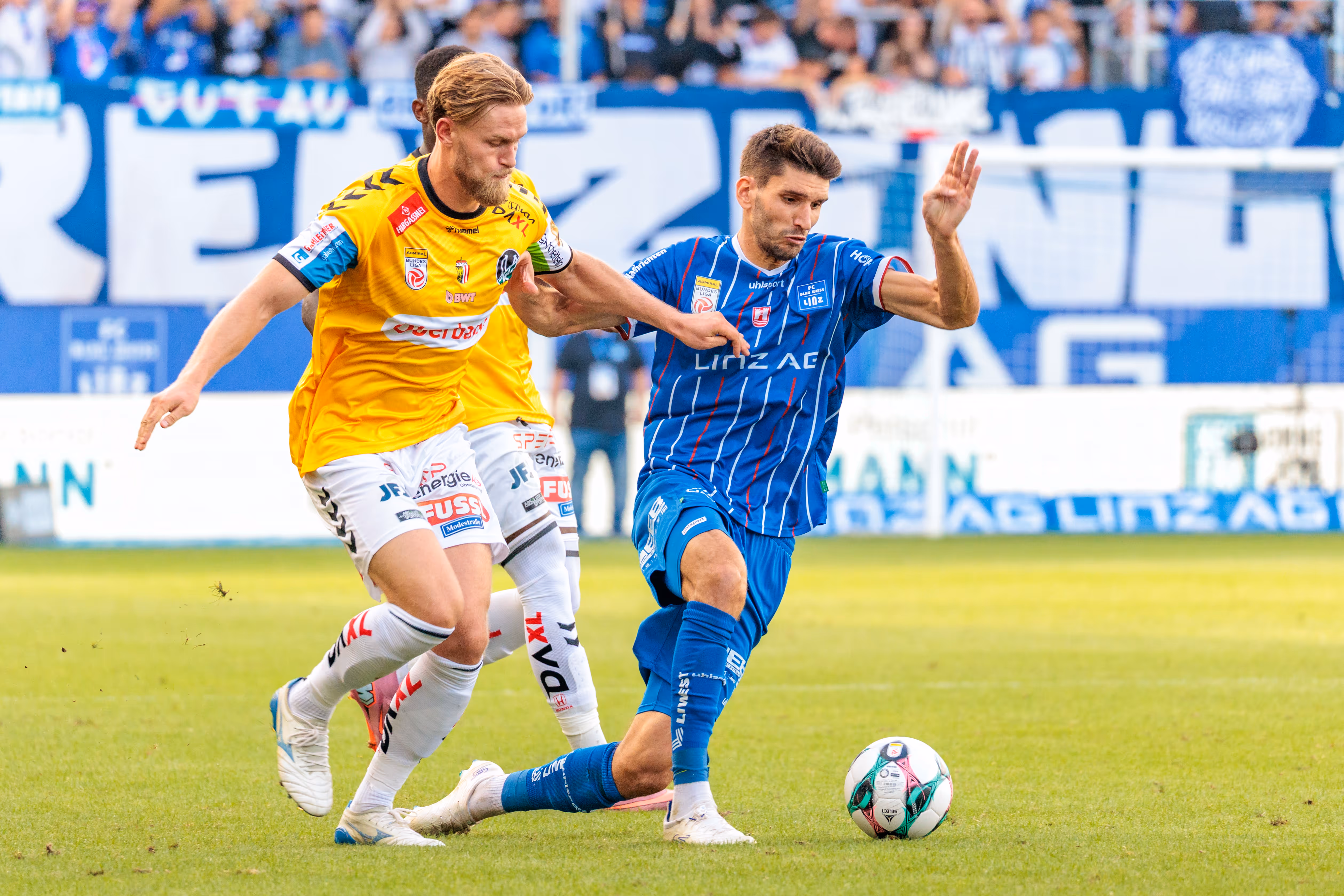 Ein Spieler des SV Ried im gelben Trikot setzt einen Spieler des FC Blau-Weiß Linz im blau-weiß gestreiften Trikot im Zweikampf um den Ball unter Druck