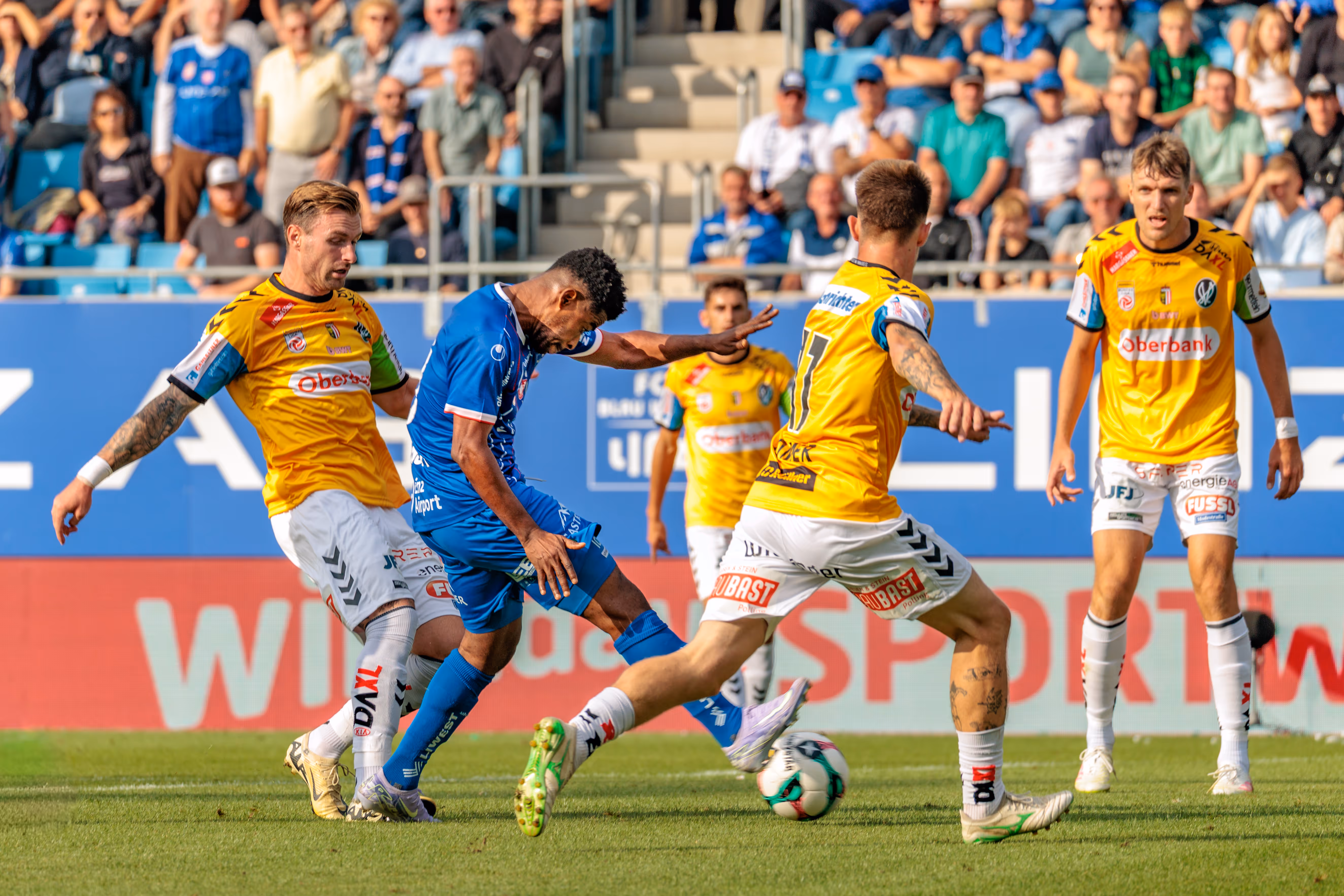 Intensiver Zweikampf um den Ball zwischen Fußballspielern auf dem Spielfeld, beobachtet von der Zuschauerkulisse im Stadion