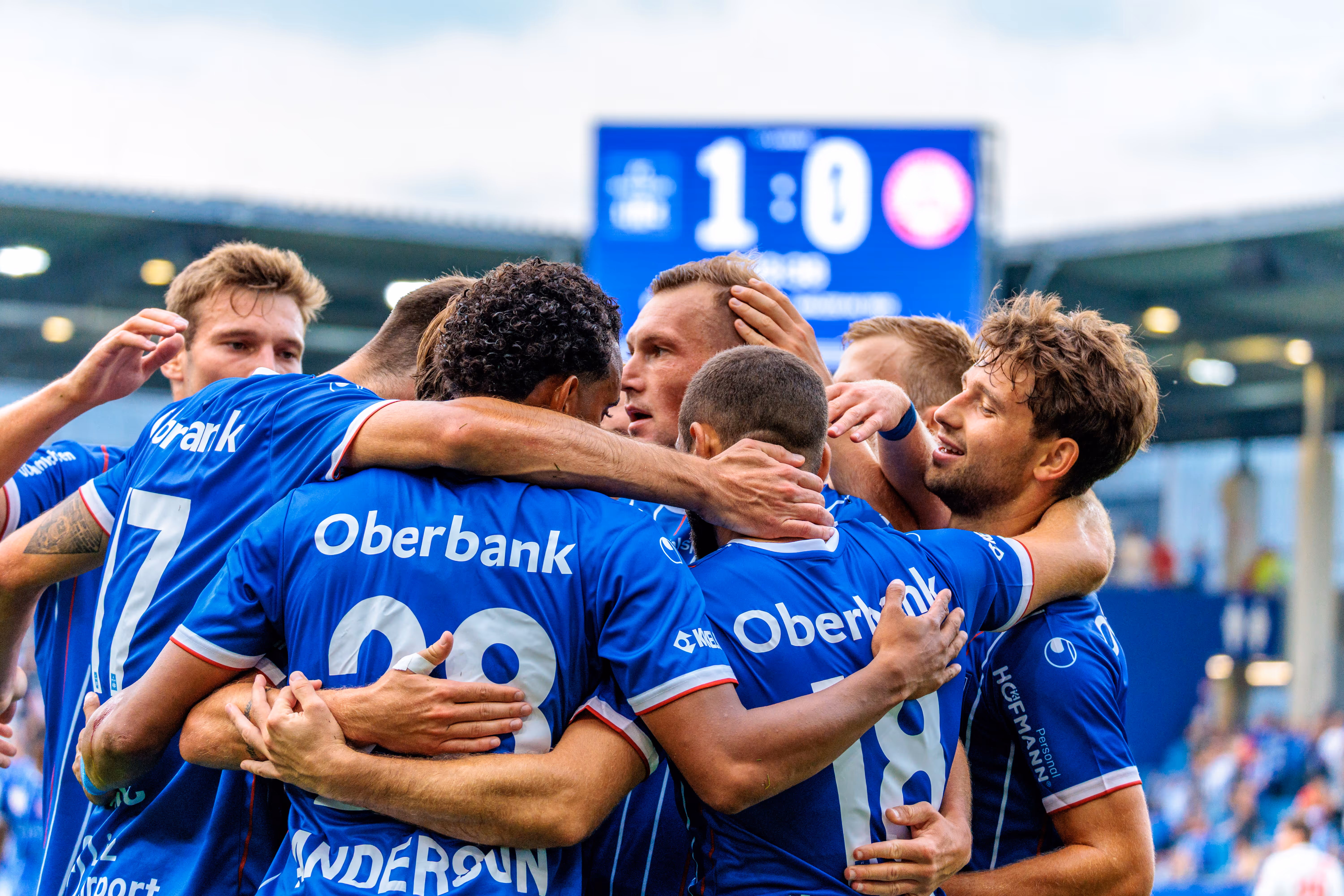 Blau-Weiß Linz Fußballspieler in blauen Trikots feiern eng umschlungen einen Führungstreffer, während die Anzeigetafel im Hintergrund das Ergebnis 1:0 anzeigt