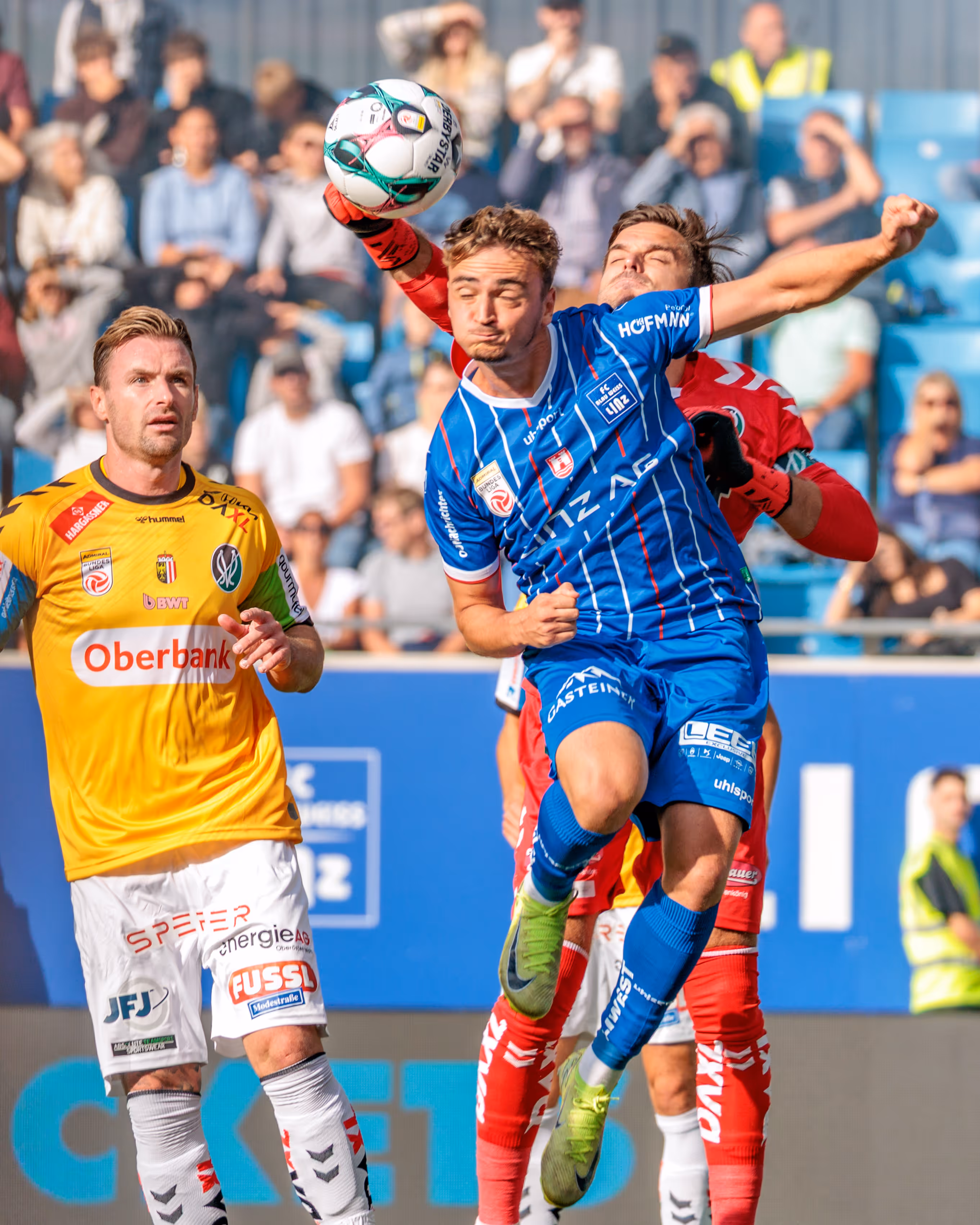 Der Torwart des SV Ried klärt den Ball mit einem Kopfball im Strafraum unter dem Druck eines Gegenspielers des Blau Weiß Linz, während weitere Spieler und Zuschauer im Hintergrund das Bundesliga-Derby verfolgen