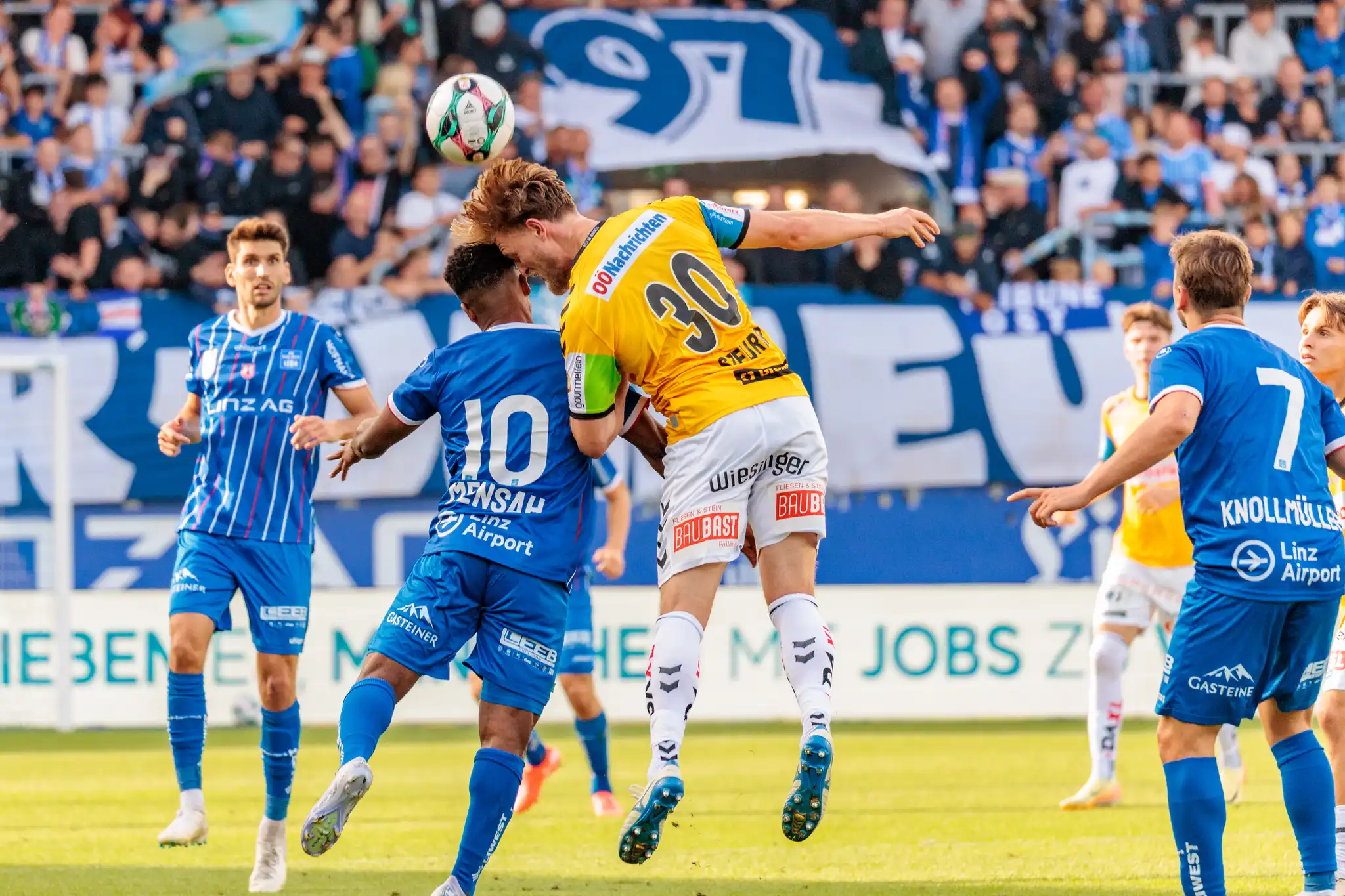 Ein Fußballer in gelbem Trikot und ein Spieler in blauem Trikot springen beim Kopfballduell um den Ball, während im Hintergrund Zuschauer auf den Tribünen zu sehen sind