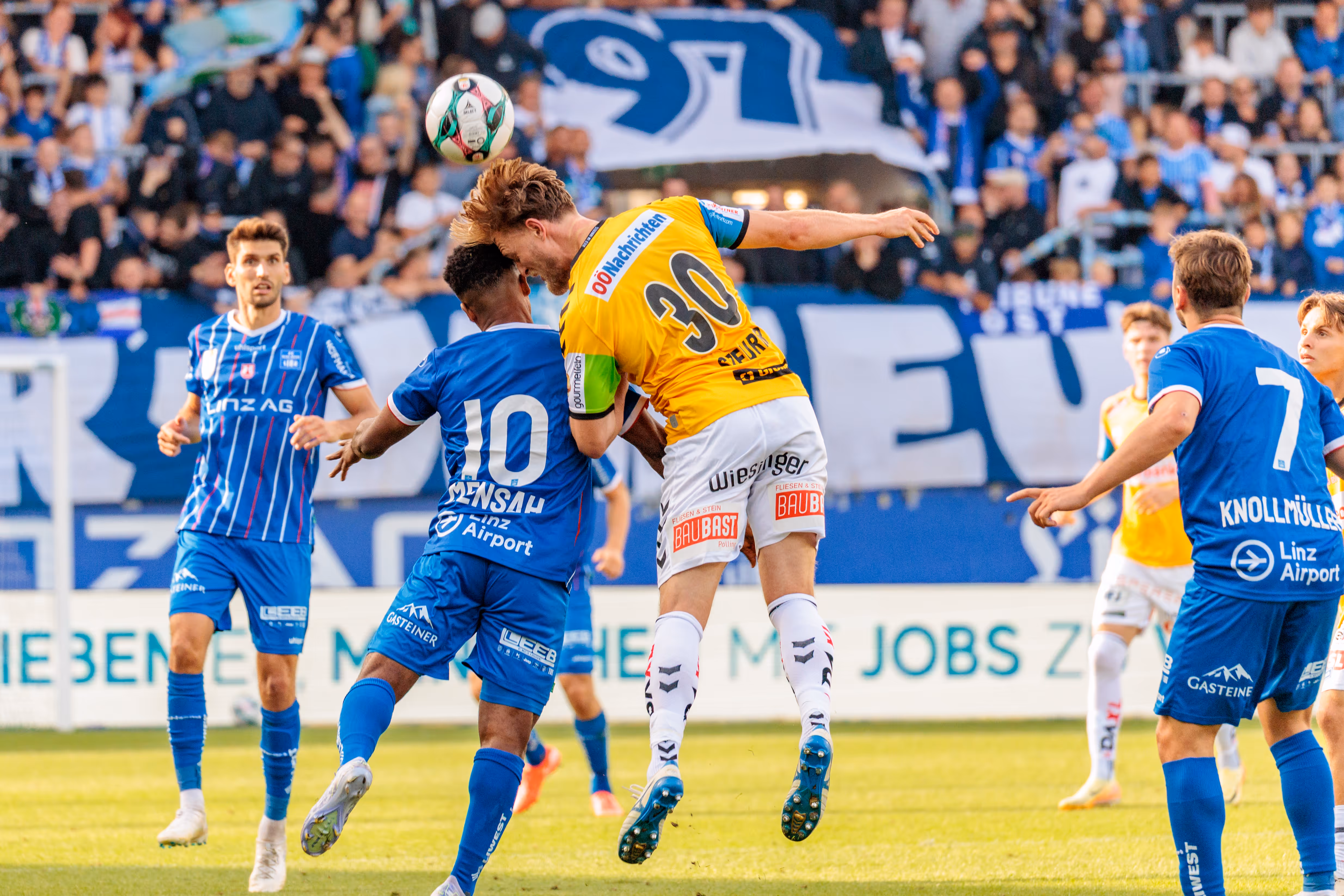 Ein Fußballer in gelbem Trikot und ein Spieler in blauem Trikot springen beim Kopfballduell um den Ball, während im Hintergrund Zuschauer auf den Tribünen zu sehen sind