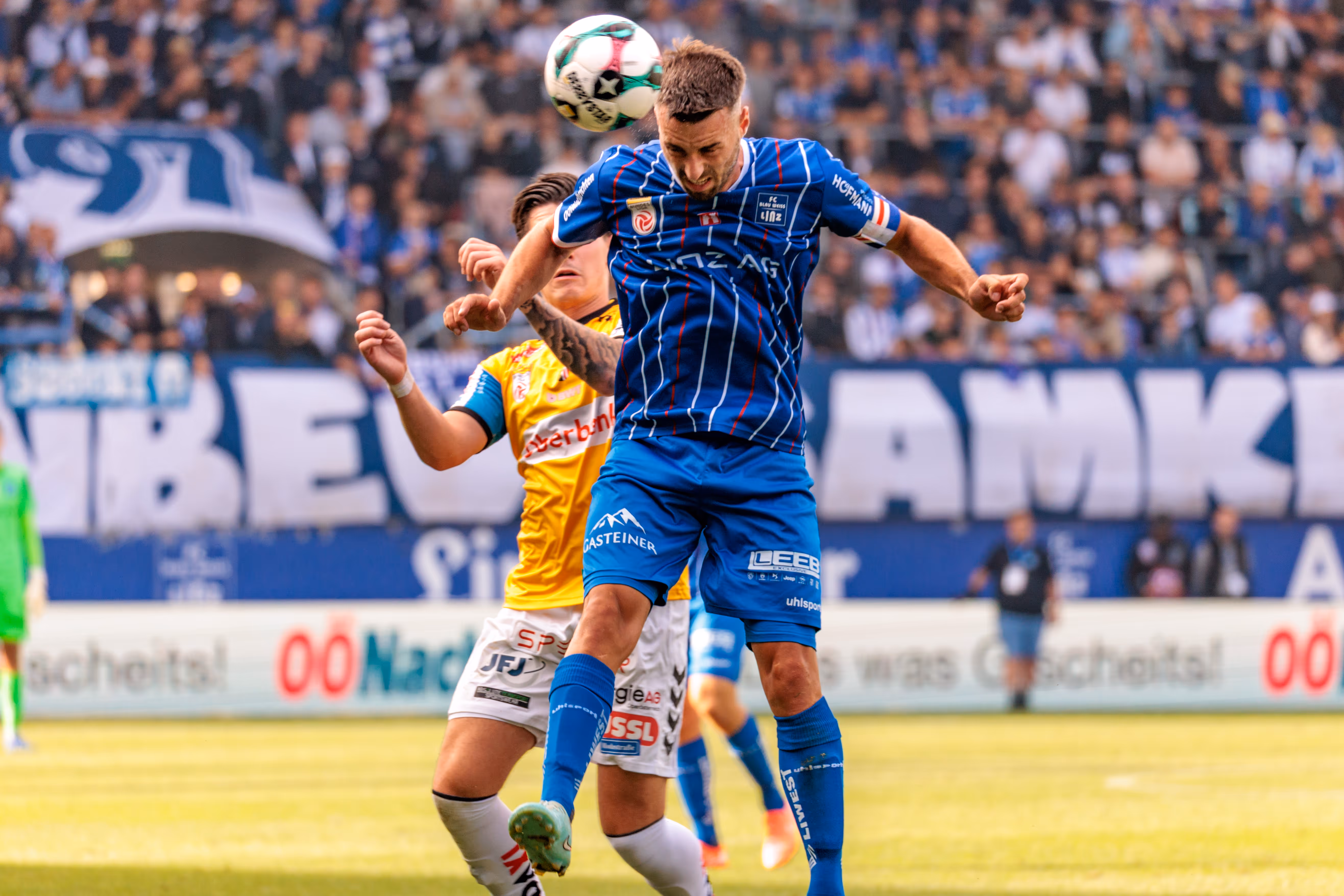 Ein Spieler des FC Blau-Weiß Linz im blauen Trikot und ein Spieler des SV Ried im gelben Trikot liefern sich ein dynamisches Kopfballduell um den Fußball