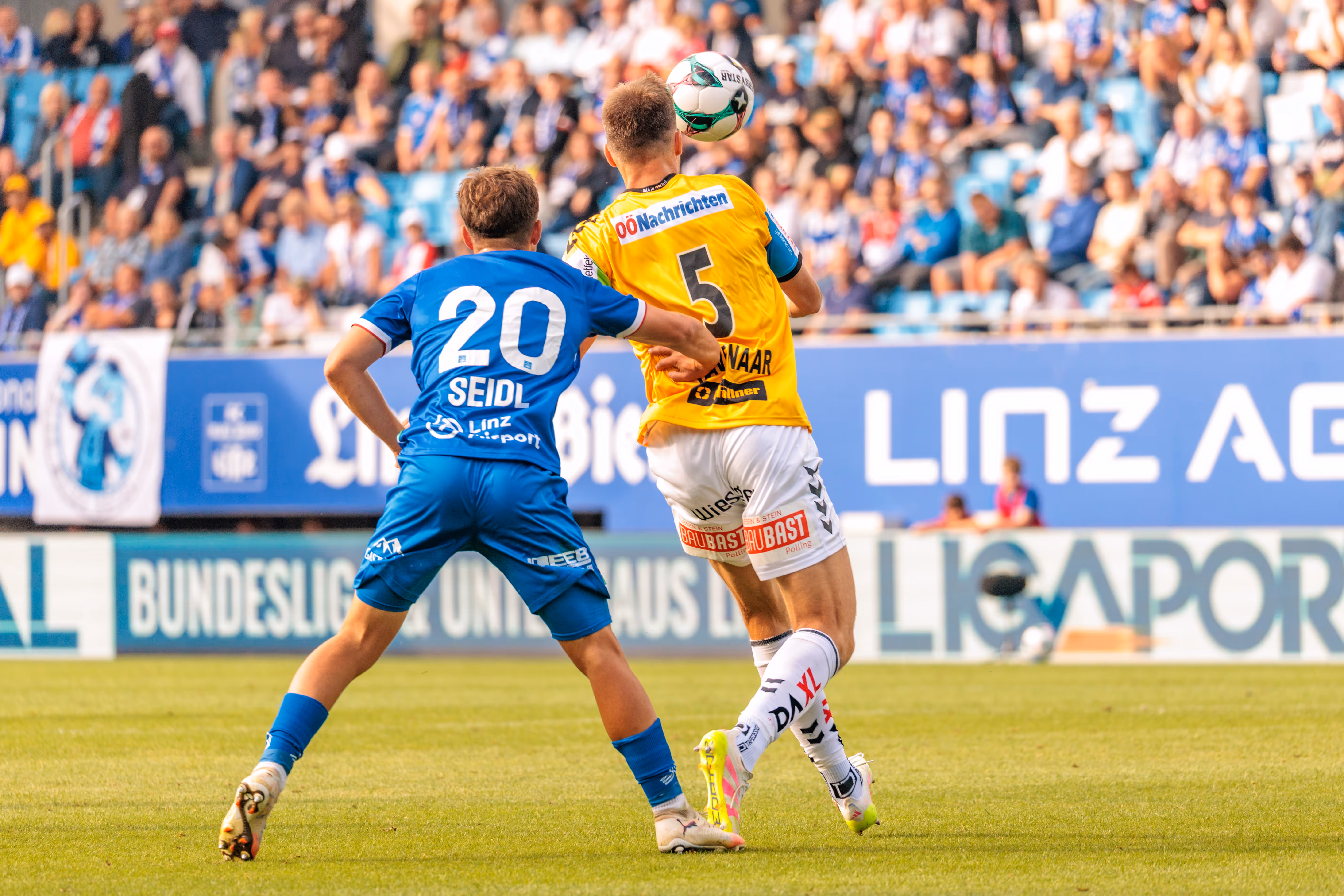Ein Fußballspieler im gelben Trikot köpft den Ball in einem Duell mit einem Spieler im blauen Trikot, beobachtet von einer verschwommenen Zuschauermenge auf der Tribüne