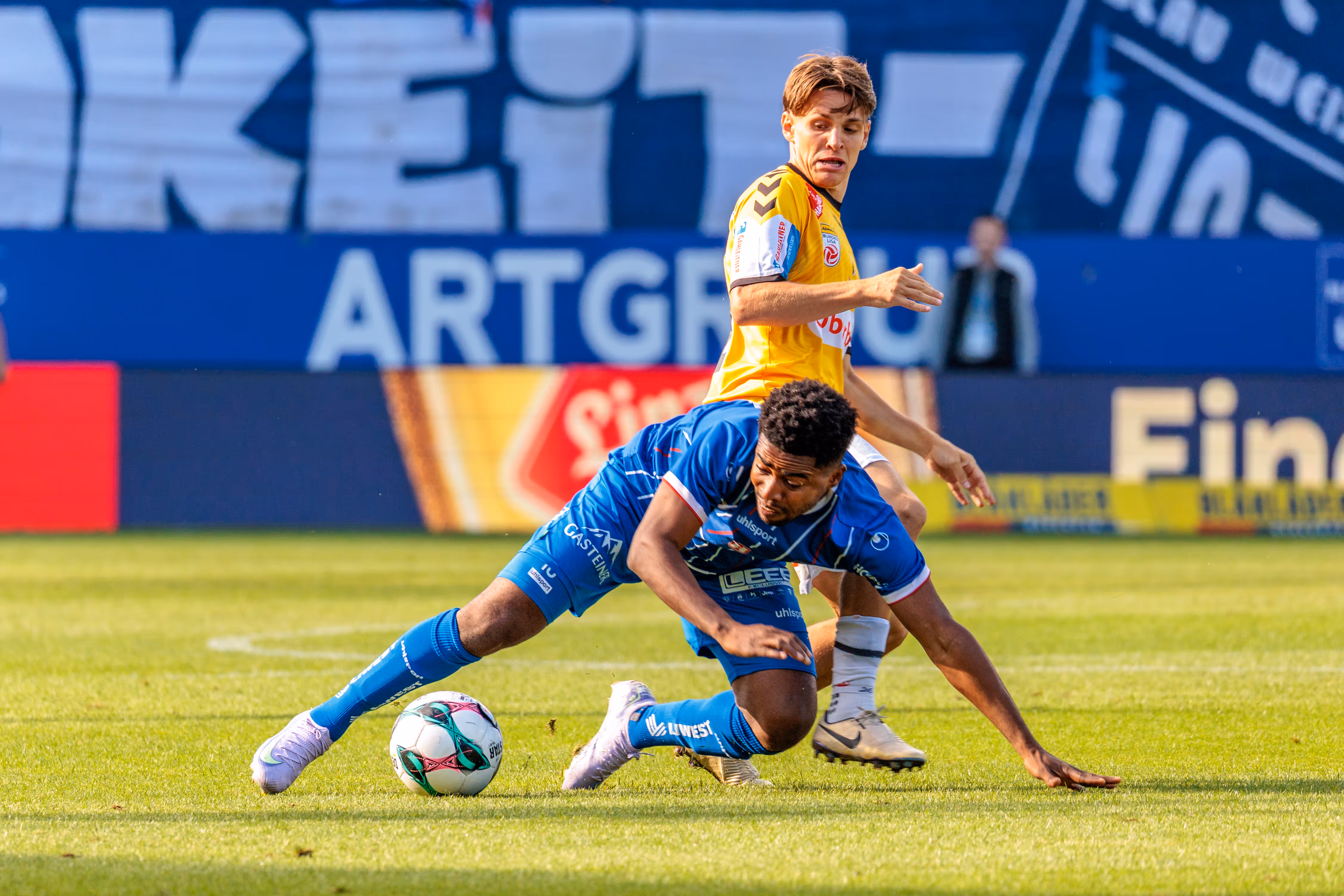 Fußball-Zweikampf im Bundesliga-Derby: Ein Spieler von Blau-Weiß Linz im blauen Trikot geht zu Boden, um den Ball vor einem Gegenspieler von SV Ried im gelben Trikot zu behaupten