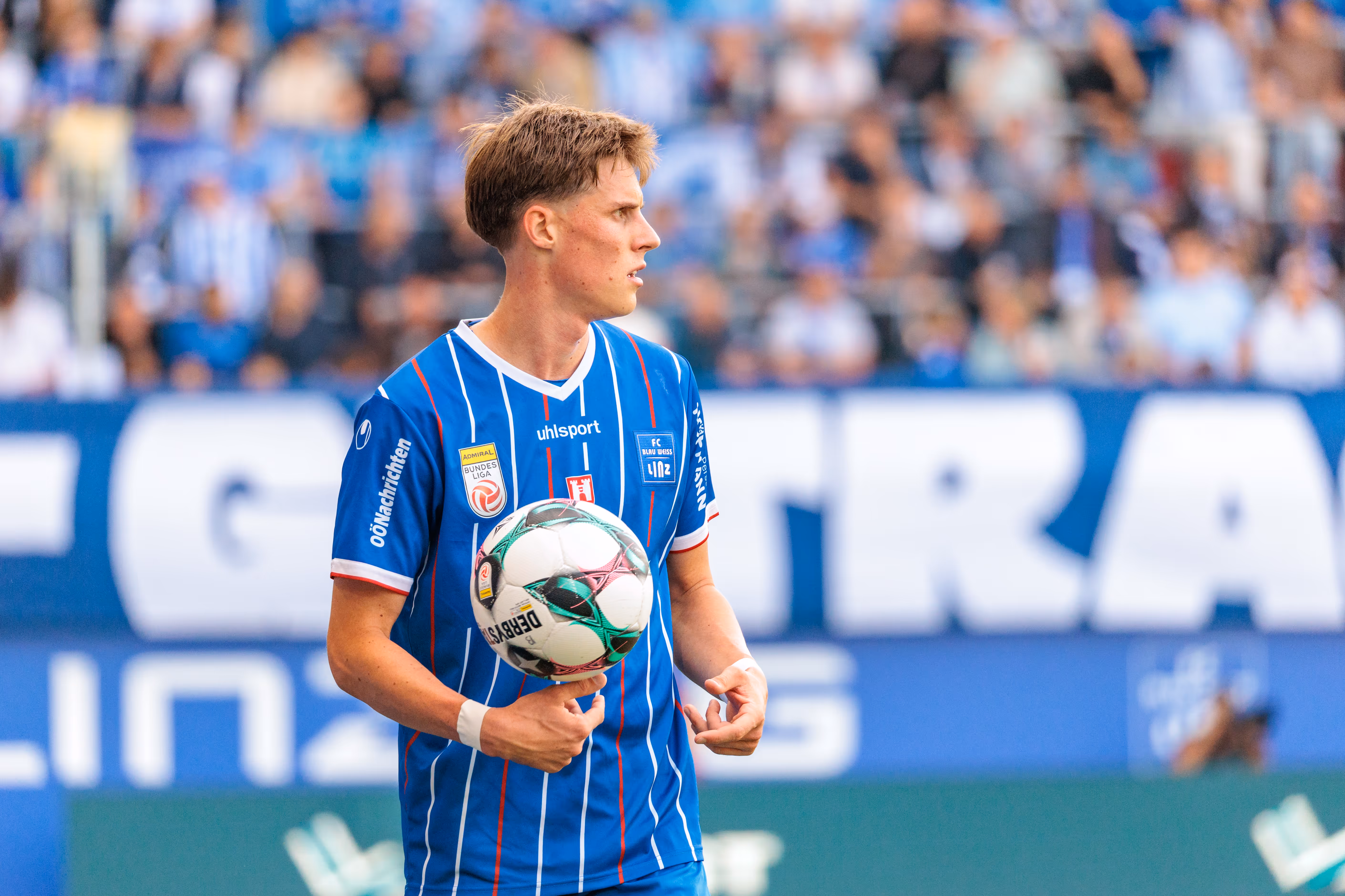 Porträt eines konzentrierten Fußballspielers des FC Blau-Weiß Linz in blau-weißem Trikot, der einen Ball im Stadion hält