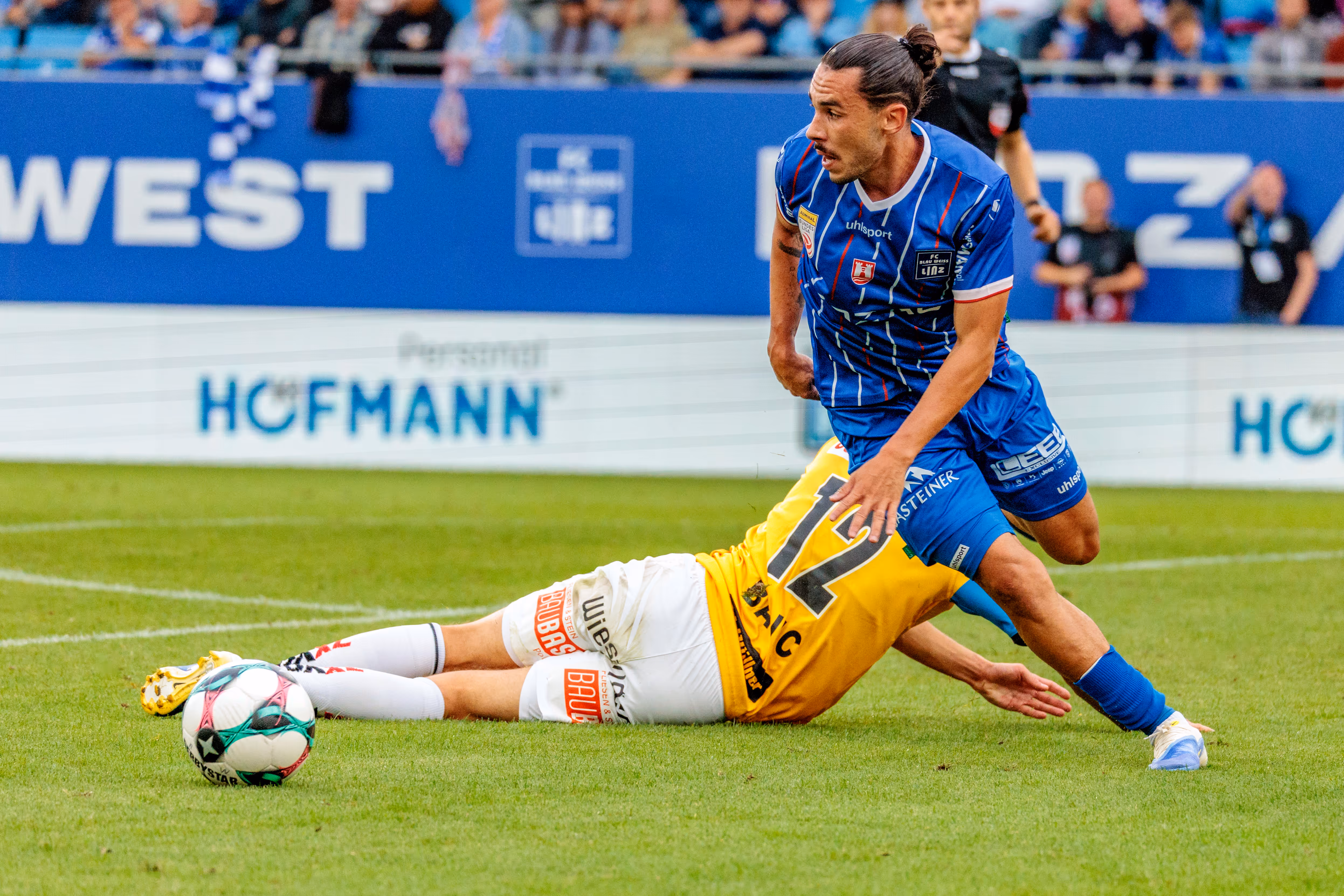 Ein Spieler von Blau-Weiß Linz steht über einem am Boden liegenden Gegner von SV Ried nach einem intensiven Zweikampf, der Fußball liegt in der Nähe