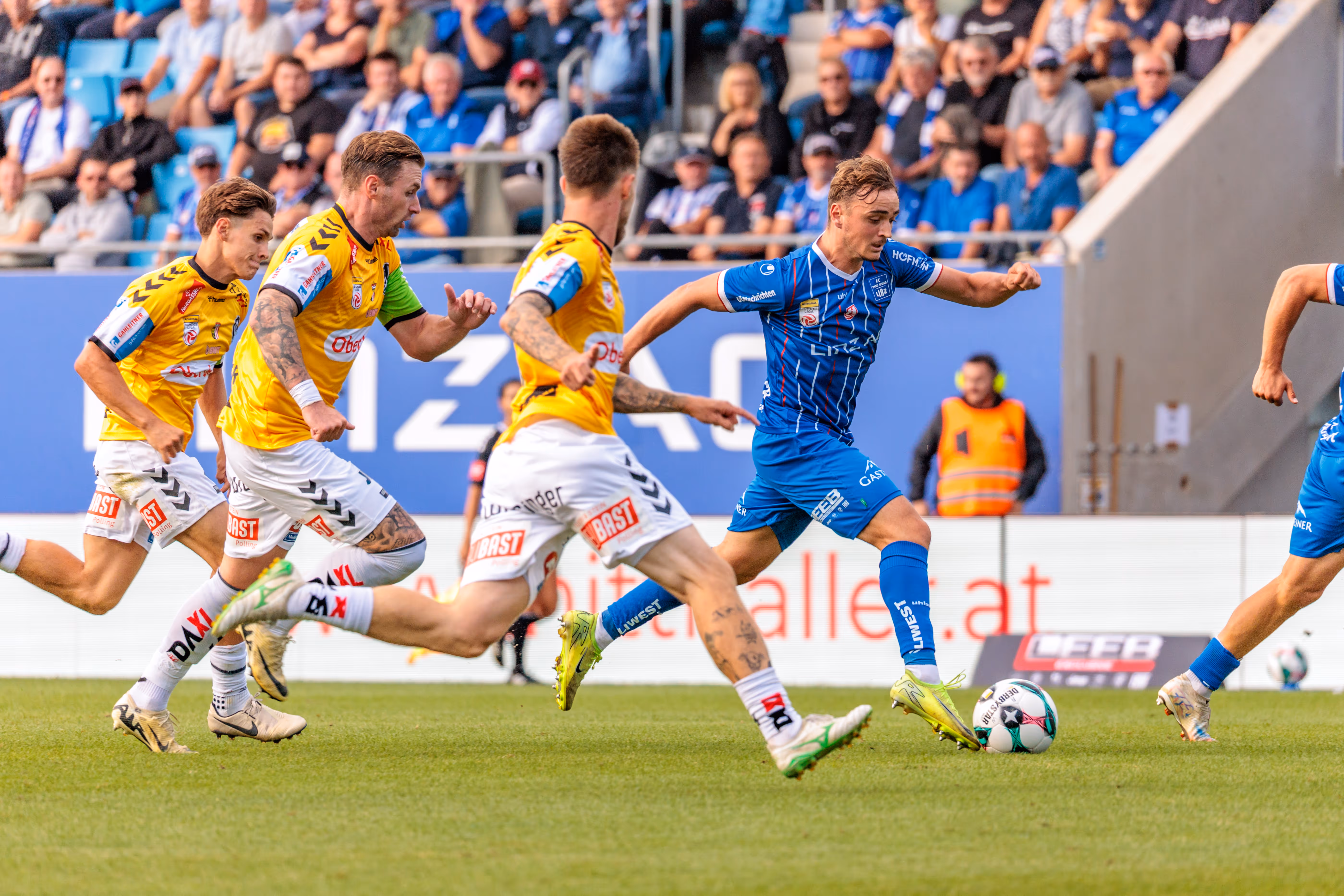 Ein Spieler des FC Blau-Weiß Linz im blauen Trikot versucht einen Torschuss auf dem Fußballfeld, während drei Spieler der SV Ried in gelben Trikots verteidigen