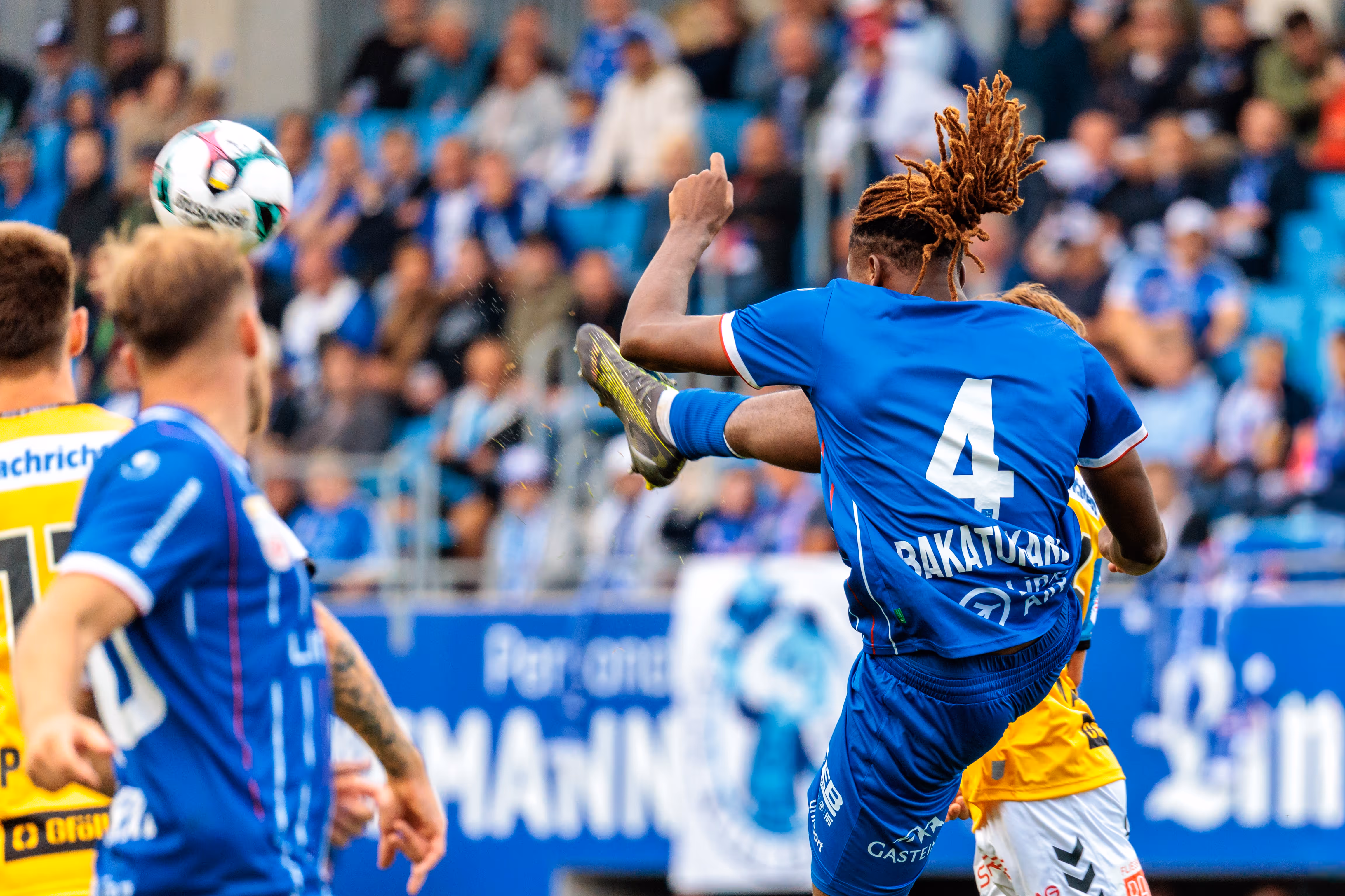Ein Verteidiger von Blau-Weiß Linz mit der Rückennummer 4 und Dreadlocks klärt den Fußball mit einem kräftigen Schuss im Spiel gegen SV Ried vor der unscharfen Kulisse der Zuschauer