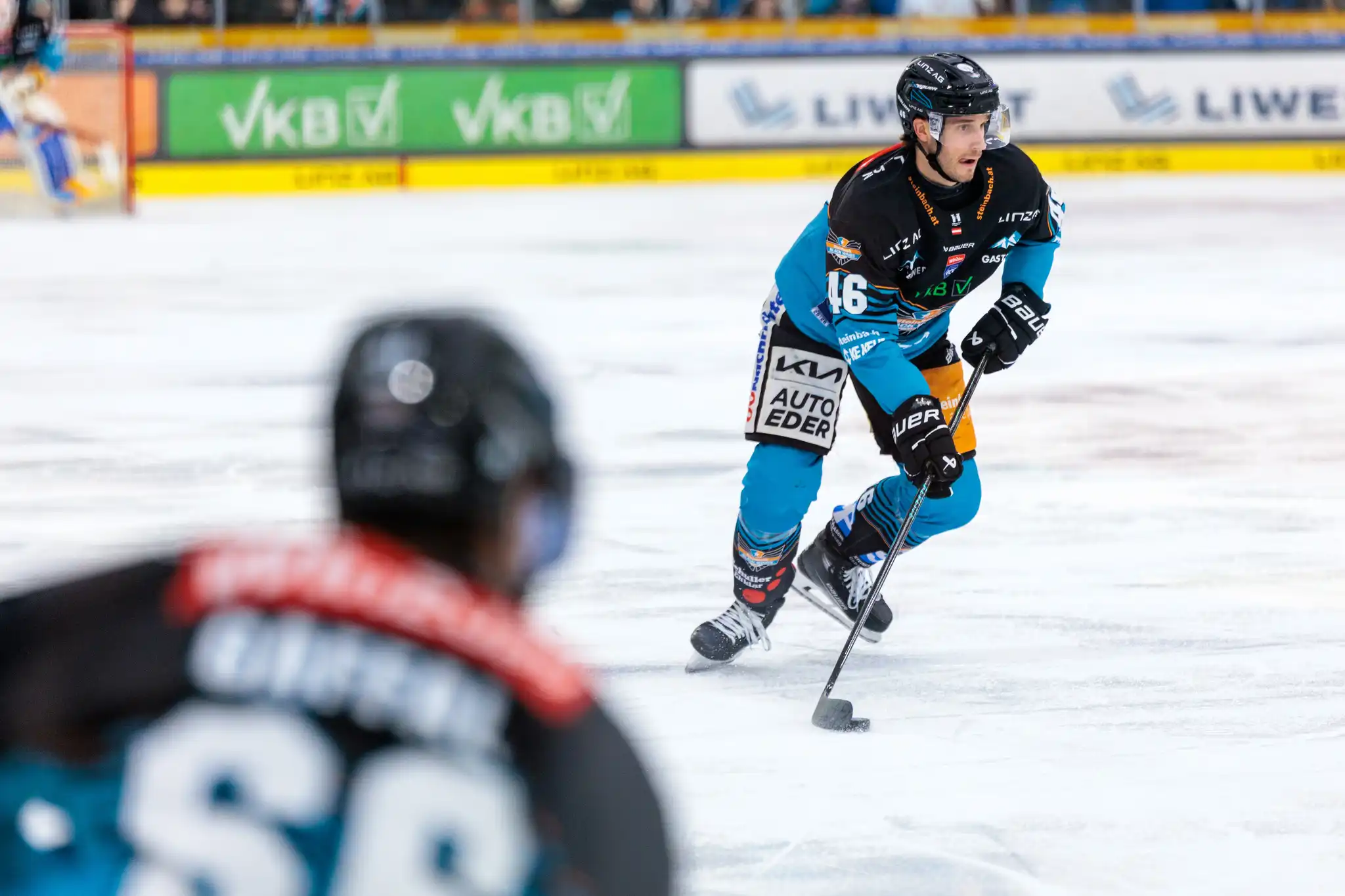 Ein Eishockeyspieler der Black Wings Linz in schwarzer und türkiser Uniform fährt auf dem Eis und hat den Puck am Schläger