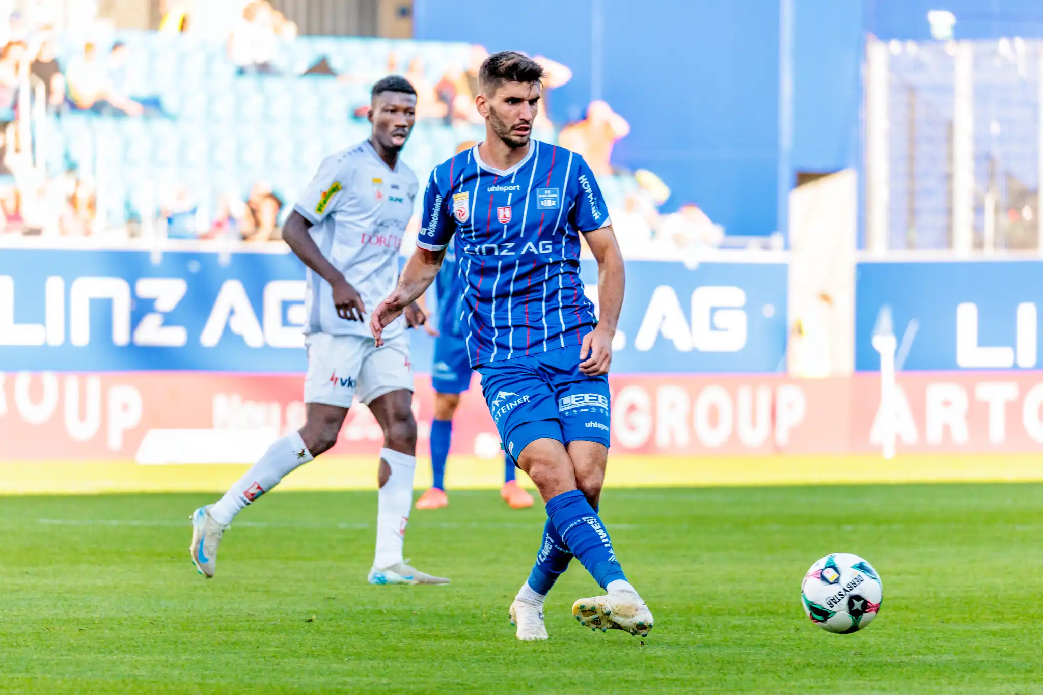 Der Mittelfeldspieler des FC Blau-Weiß Linz, Christopher Cvetko, kontrolliert in einer dynamischen Spielszene den Fußball, während ihn ein Gegenspieler im Hintergrund beobachtet