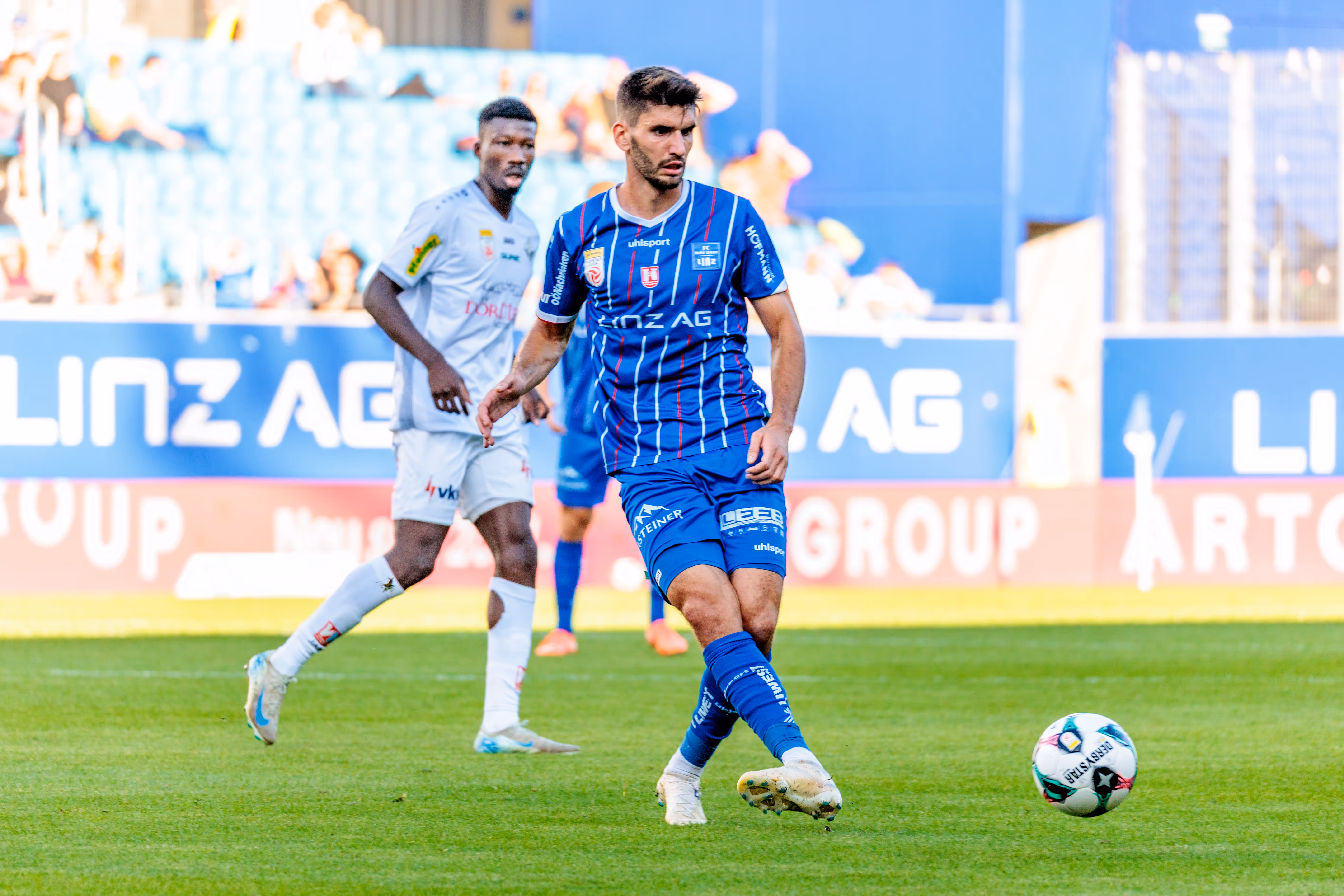 Der Mittelfeldspieler des FC Blau-Weiß Linz, Christopher Cvetko, kontrolliert in einer dynamischen Spielszene den Fußball, während ihn ein Gegenspieler im Hintergrund beobachtet