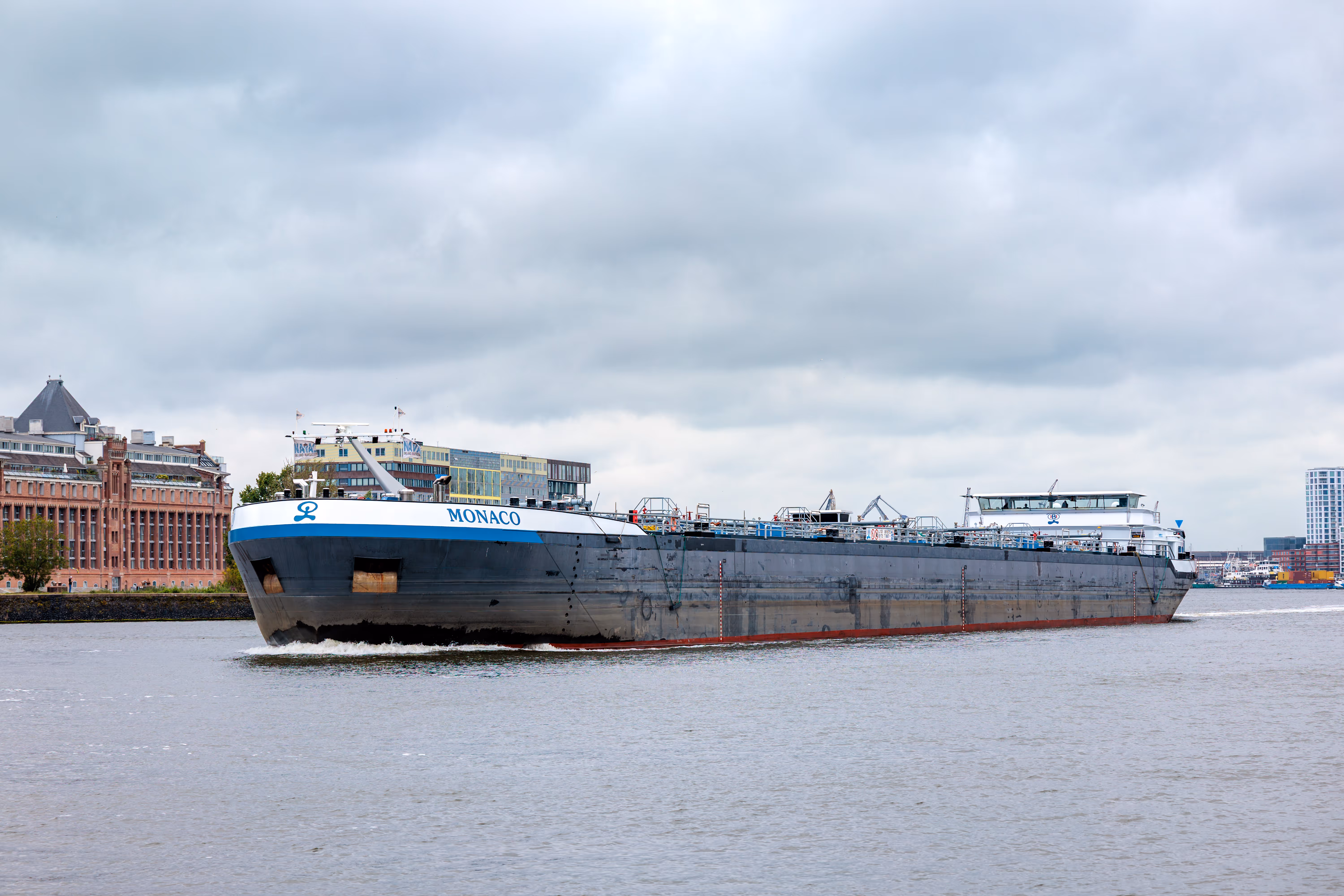 Ein großes, graues Frachtschiff namens Monaco befährt unter einem bewölkten Himmel einen Wasserweg in Amsterdam, gesäumt von verschiedenen Stadtgebäuden - 2025/06/10