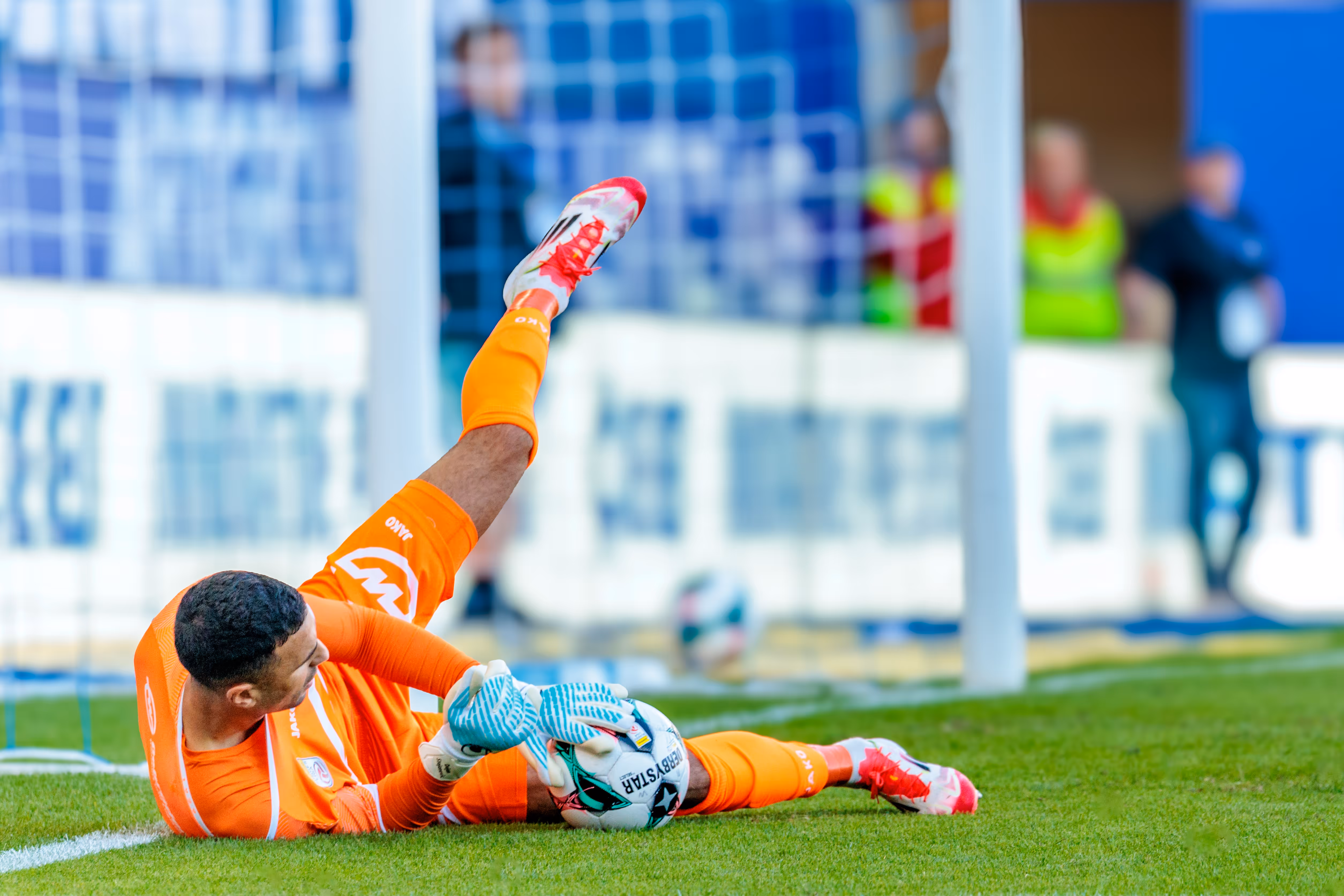 Der Tormann des SCR Altach, Dejan Stojanovic, zeigt in seinem leuchtend orangen Trikot eine spektakuläre Parade, bei der er sich voll ausstreckt und den Ball sicher am Boden festhält