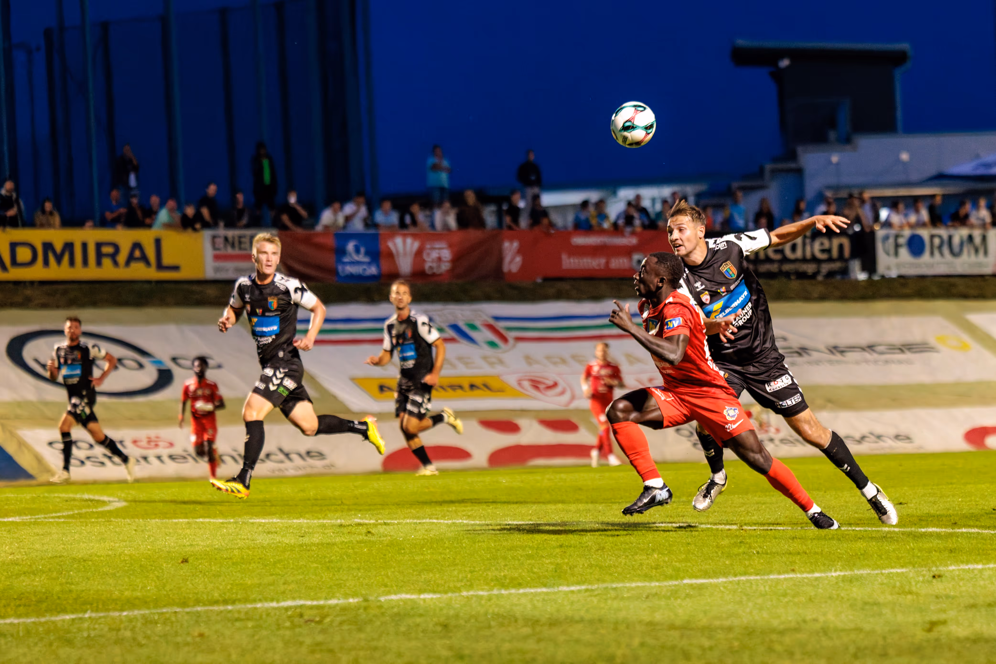 Zwei Fußballspieler, einer in rotem und einer in schwarzem Trikot, duellieren sich im Strafraum um einen hoch in der Luft schwebenden Ball während eines ÖFB-Cup-Spiels