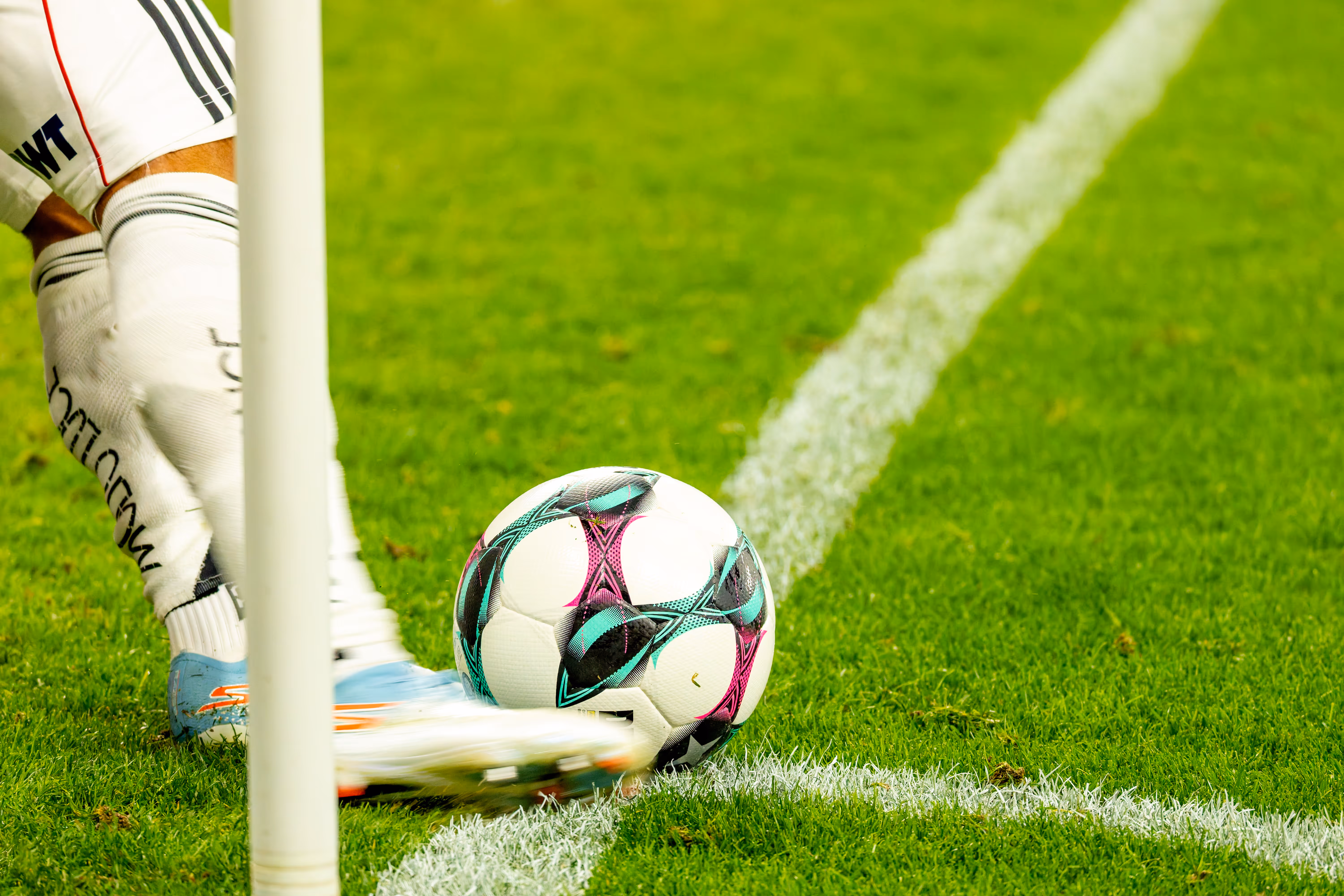 Nahaufnahme eines Fußballspielers in weißem Trikot und blauen Schuhen, dessen Fuß einen auffällig gemusterten Fußball am Eckpunkt eines Fußballfeldes in Bewegung setzt