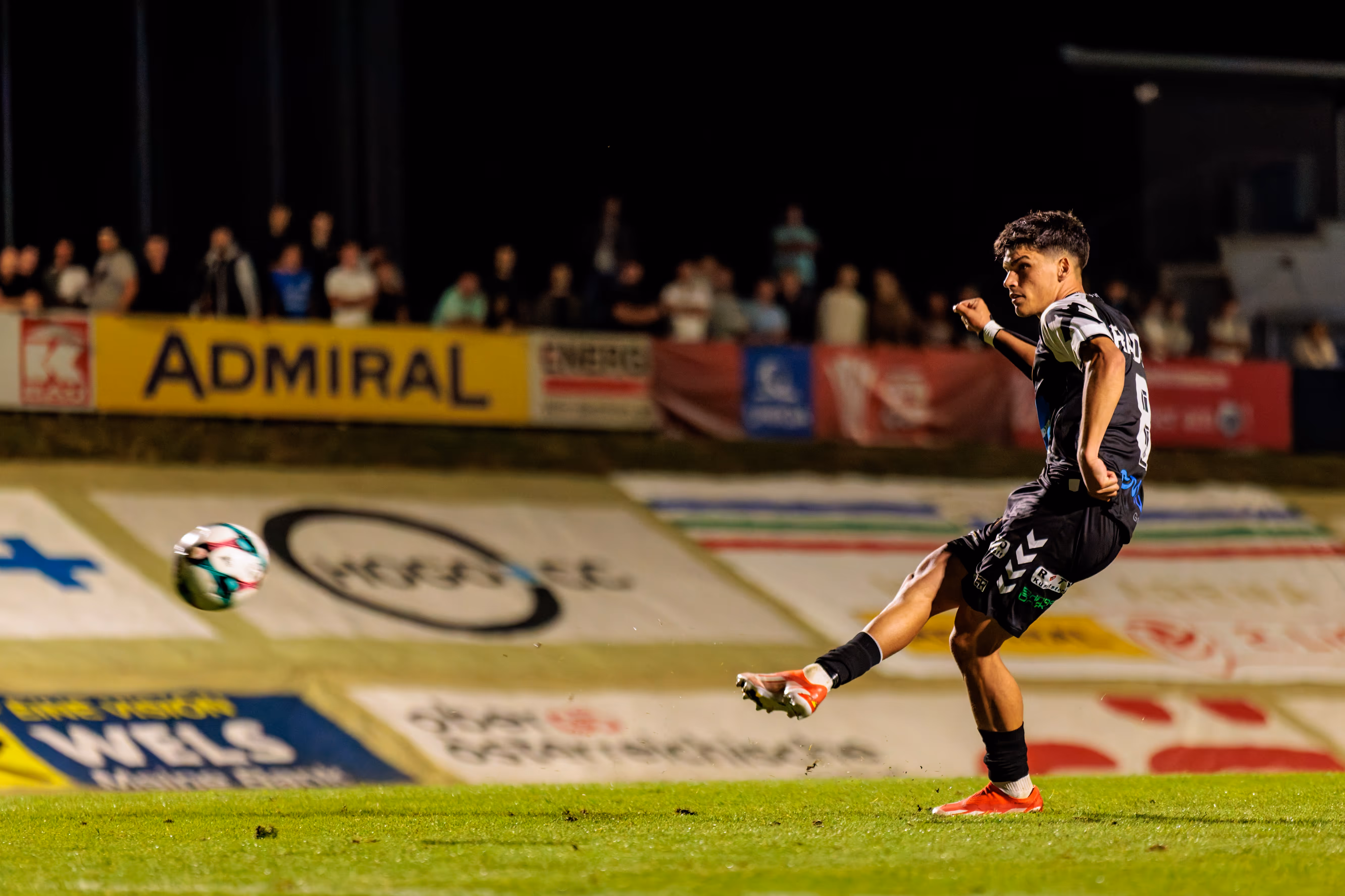 Ein Fußballspieler in schwarz-weißer Spielkleidung von SPG HOGO Wels tritt im ÖFB-Cup einen Elfmeter, wobei im Hintergrund eine Admiral-Werbebande zu sehen ist