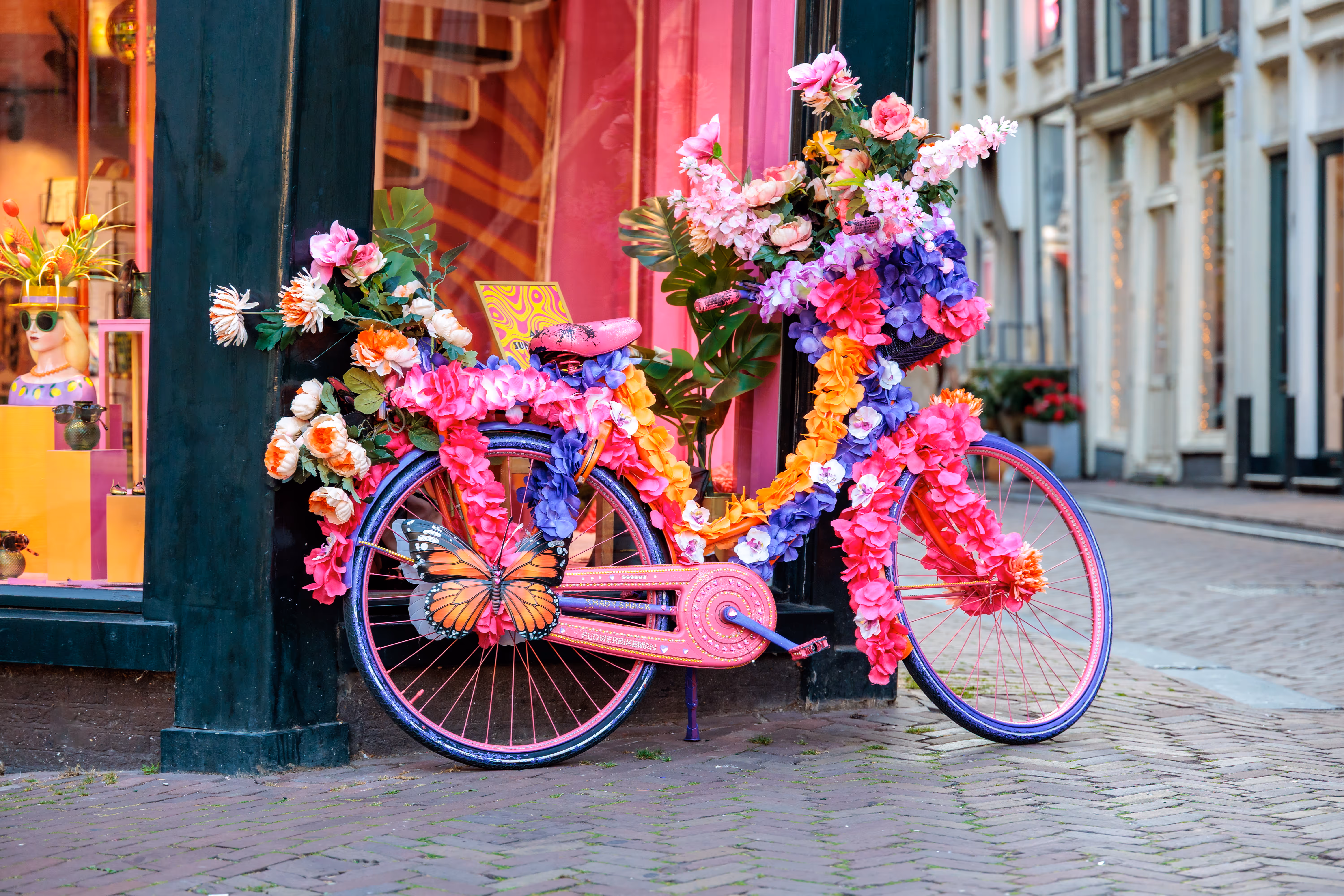 Ein farbenfrohes, mit Blumen und einem Schmetterling dekoriertes Fahrrad steht auf einem Backsteinweg in einer Amsterdamer Straße 