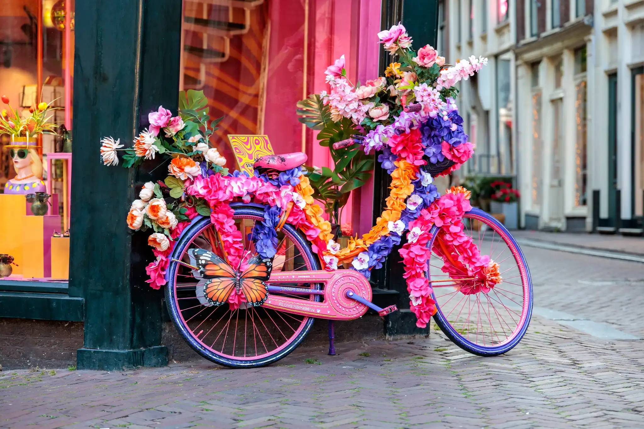 Ein farbenfrohes, mit Blumen und einem Schmetterling dekoriertes Fahrrad steht auf einem Backsteinweg in einer Amsterdamer Straße