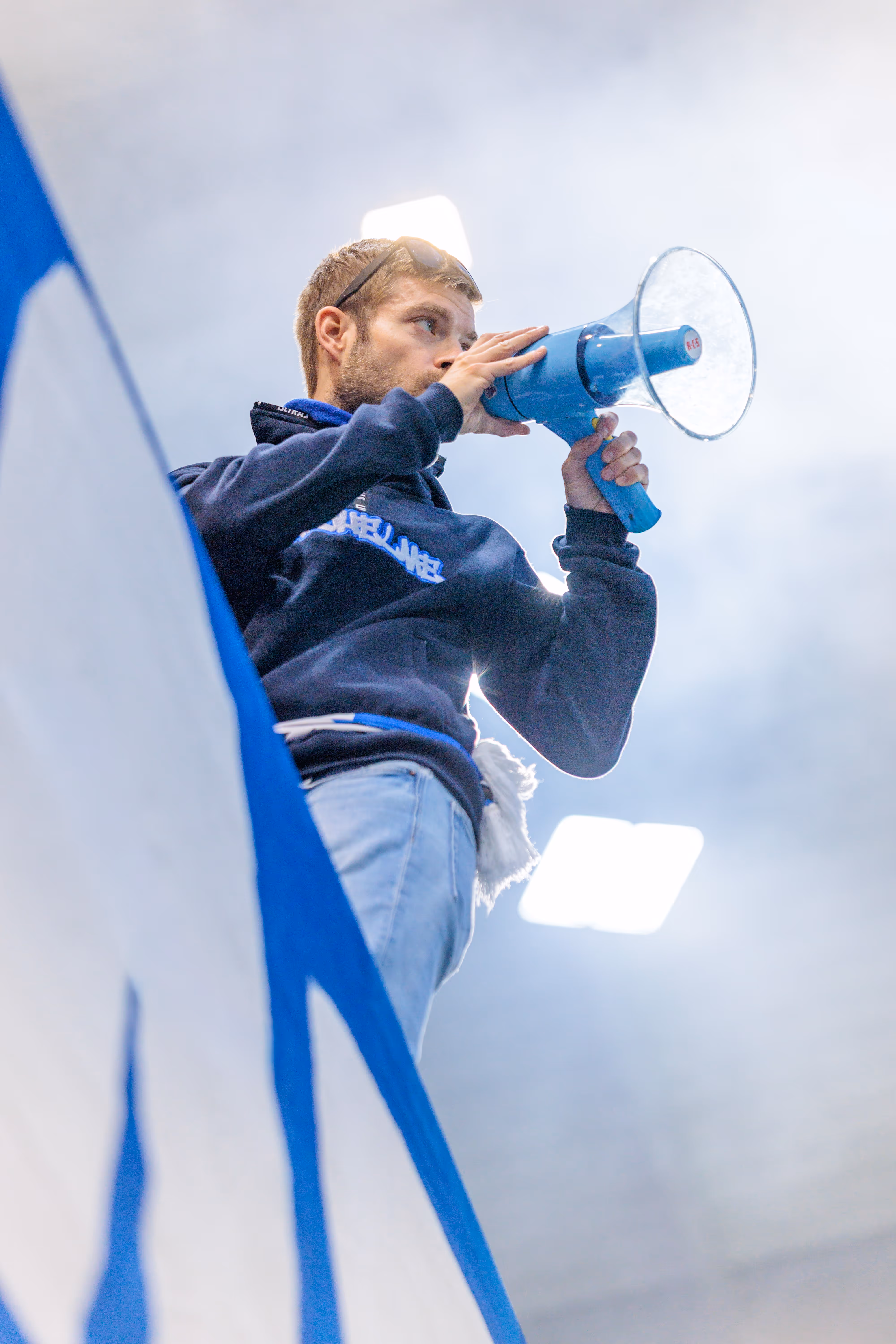 Ein junger Vorsänger in einem dunklen Kapuzenpullover heizt mit einem blauen Megaphon die Stimmung im Stadion an, fotografiert aus der Froschperspektive