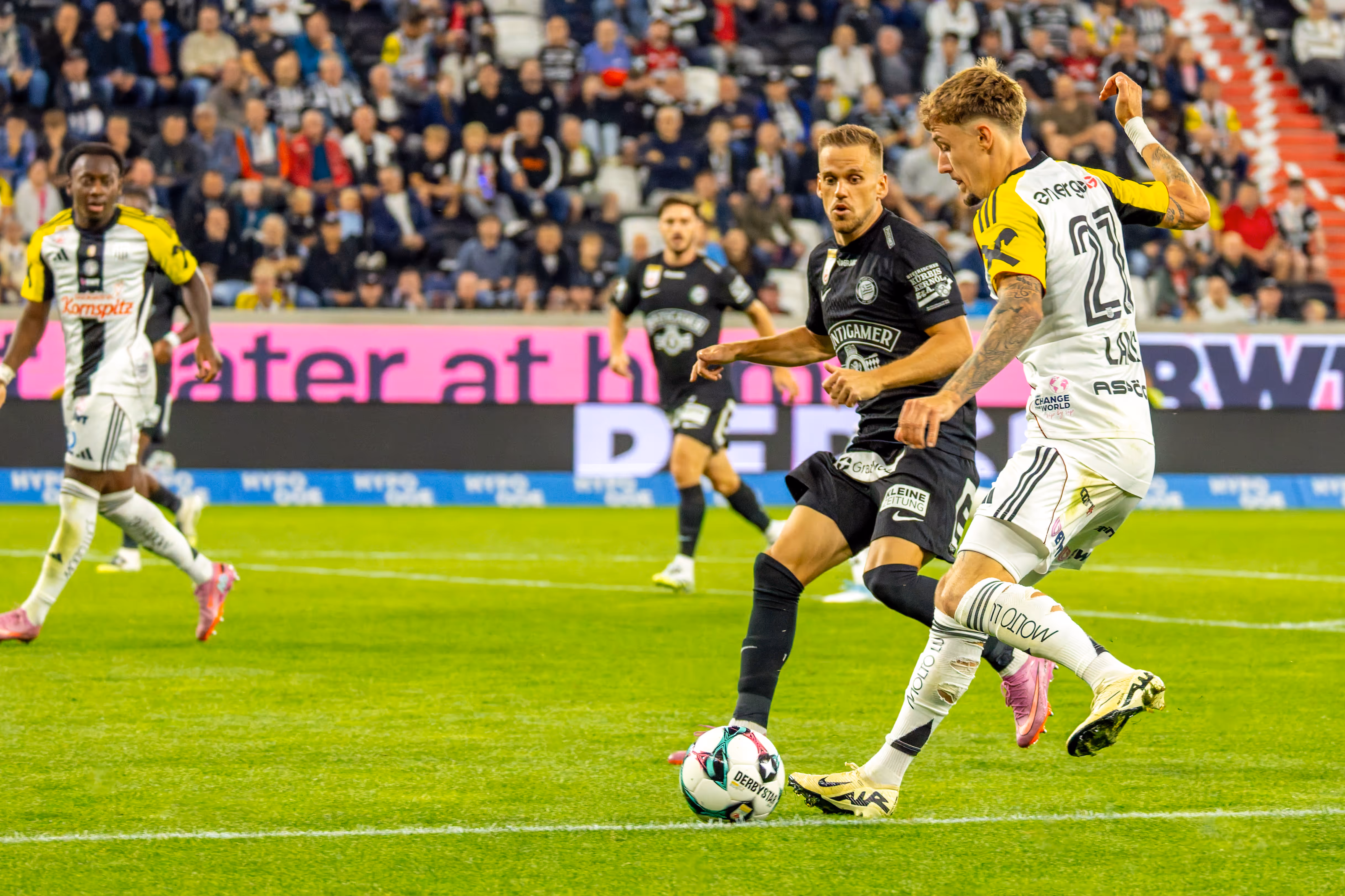 Zwei Fußballspieler, einer im hellen LASK-Trikot und einer im dunklen Trikot, ringen auf dem Spielfeld um den Ball vor einer Zuschauerkulisse