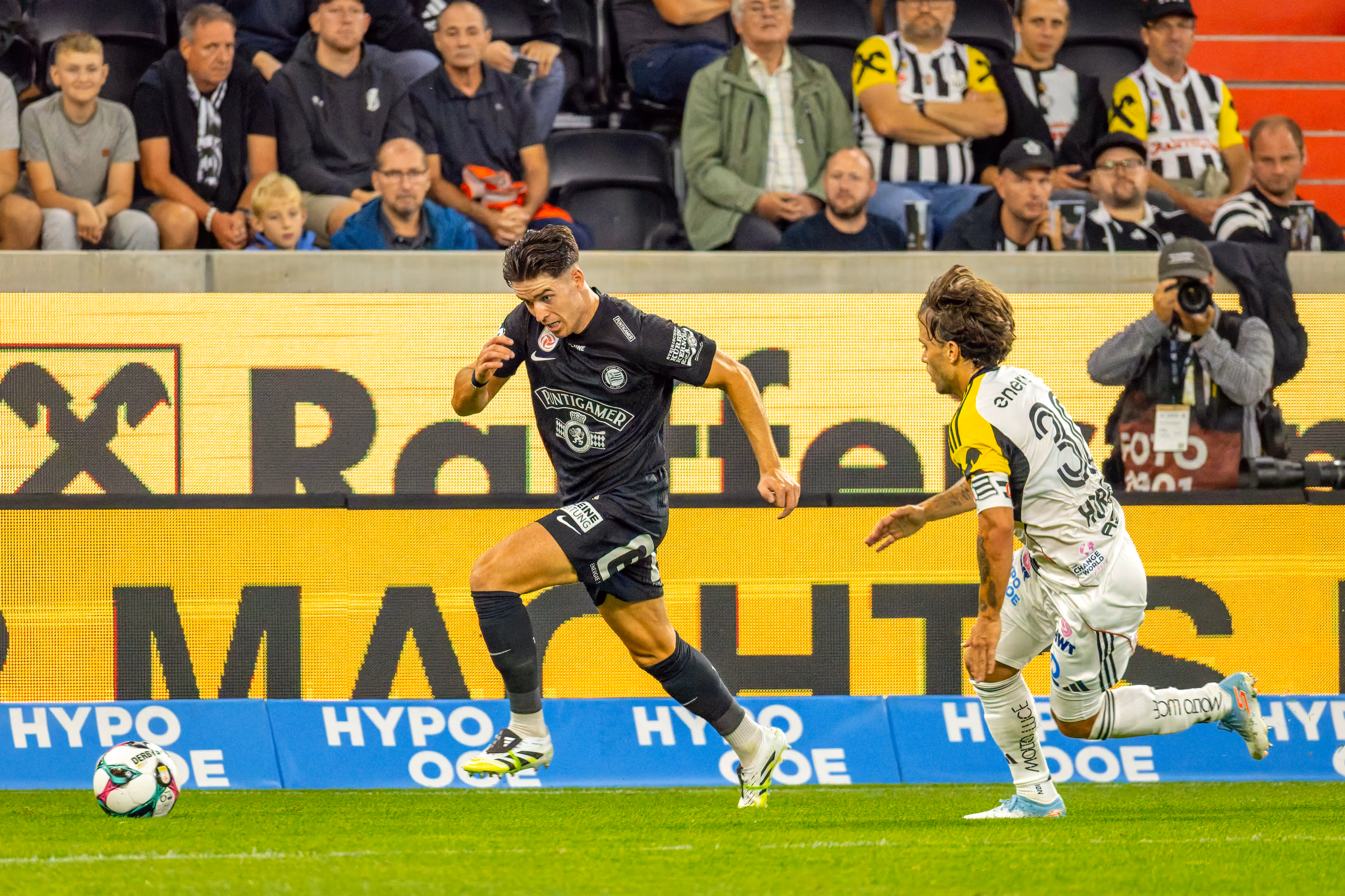Ein Fußballspieler im schwarzen Trikot rennt dynamisch mit dem Ball auf dem Feld, verfolgt von einem Gegenspieler in hellem Trikot, mit einer großen LED-Werbewand und Zuschauern im Hintergrund