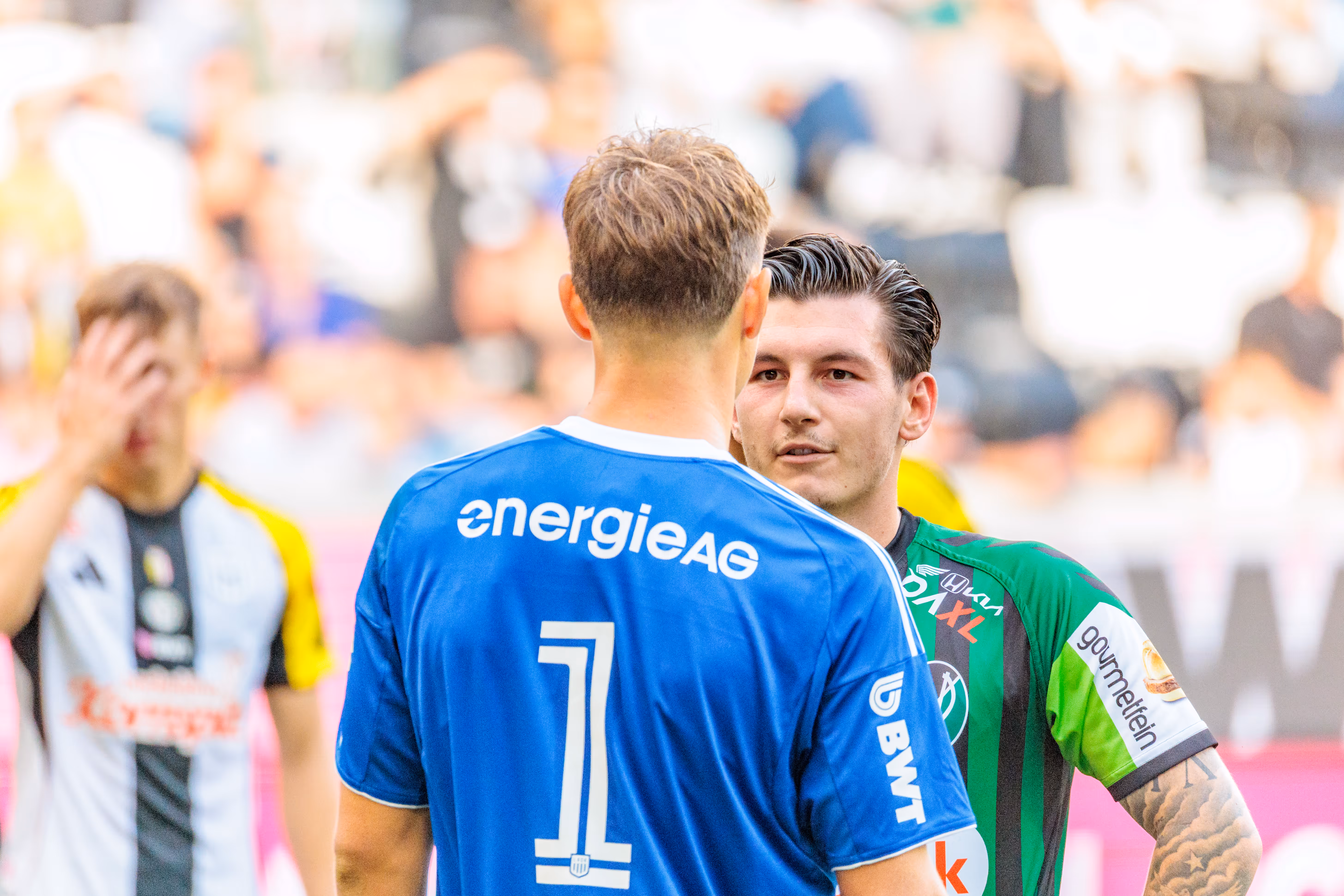Ein LASK-Torhüter im blauen Trikot mit der Nummer 1 unterhält sich mit einem SV Ried-Spieler in grün-schwarzem Trikot auf dem Fußballfeld vor der Ausführung eines Elfmeters während eines Bundesliga-Spiels