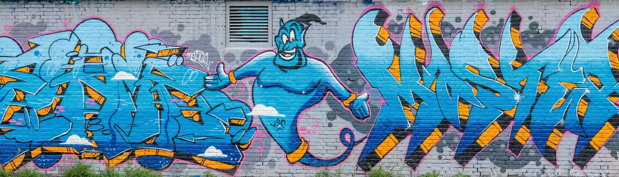 Ein farbenfrohes Graffiti-Wandbild auf einer Ziegelmauer in Amsterdam zeigt den lächelnden Dschinni aus Aladdin inmitten von stilisierten blauen und orangen Buchstaben