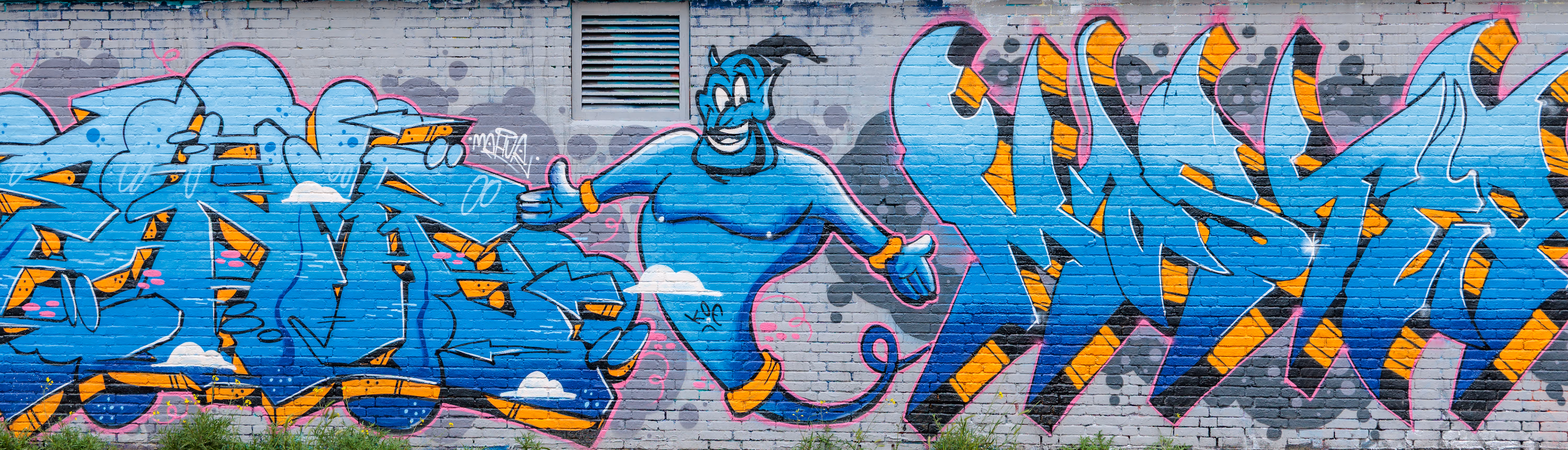 Ein farbenfrohes Graffiti-Wandbild auf einer Ziegelmauer in Amsterdam zeigt den lächelnden Dschinni aus Aladdin inmitten von stilisierten blauen und orangen Buchstaben 