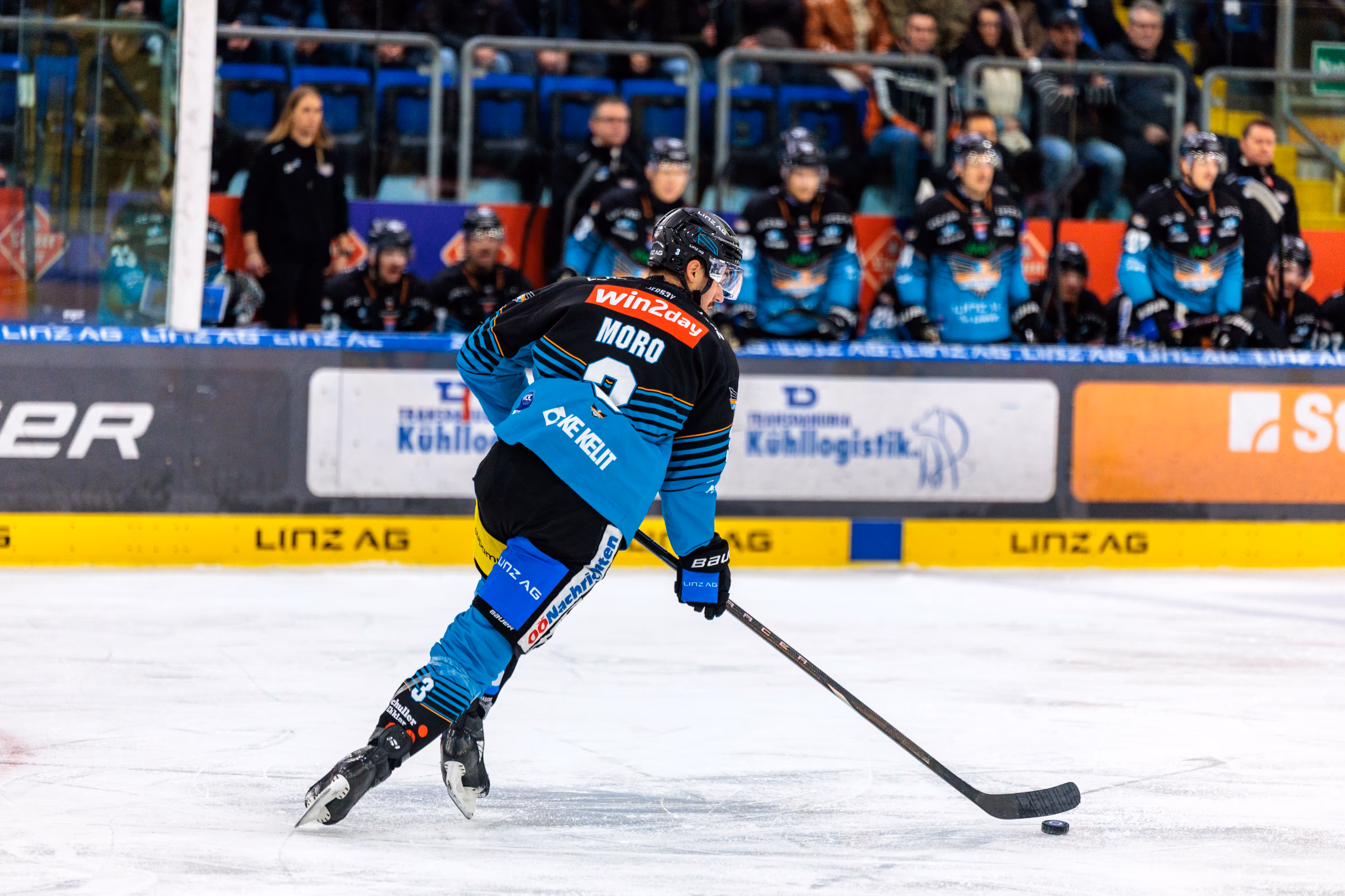 Eishockeyspieler Greg Moro (Nummer 3) im schwarzen und hellblauen Trikot skatet auf dem Eis und dribbelt den Puck mit seinem Schläger