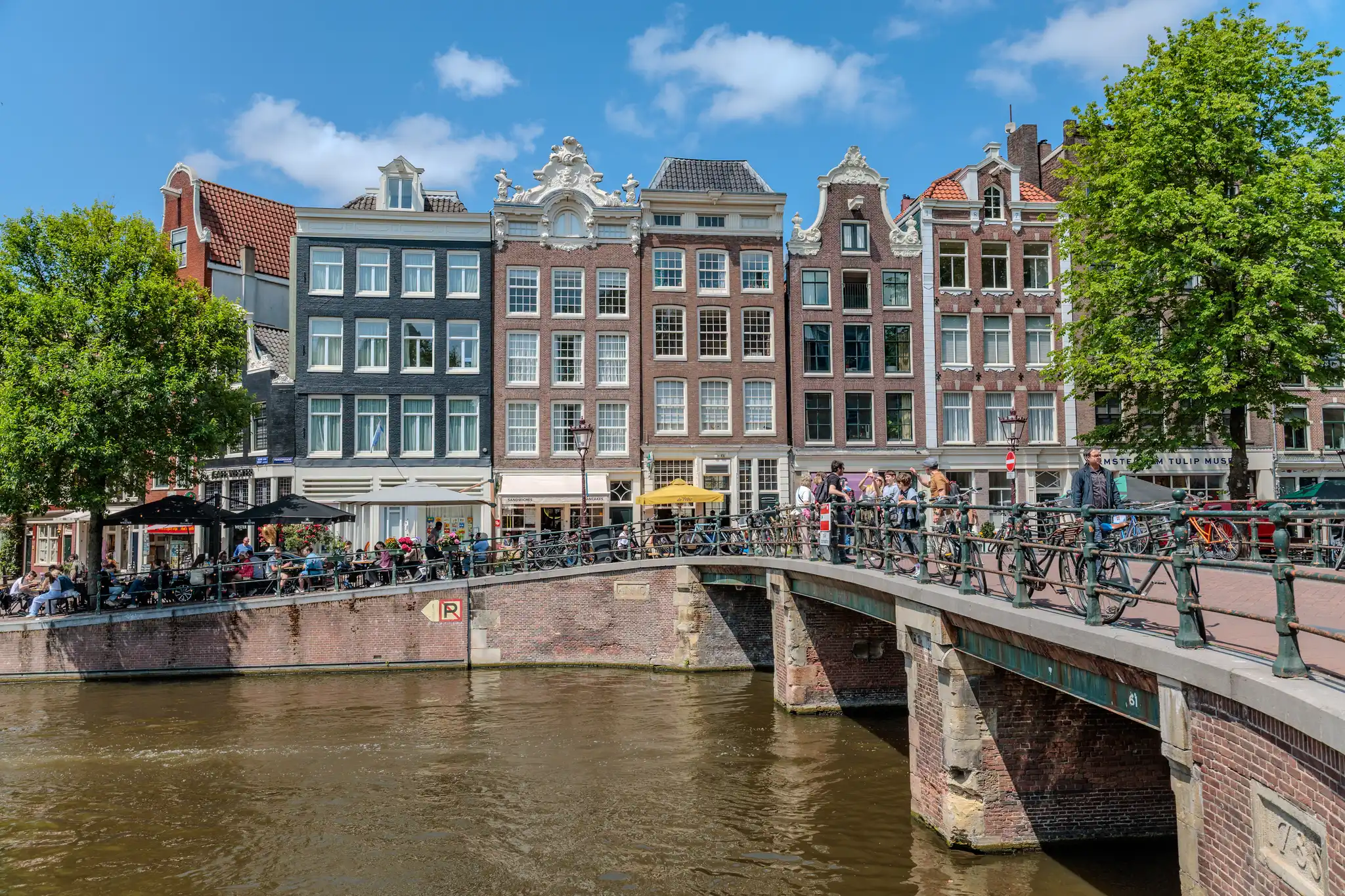 Eine malerische Szene in Amsterdam zeigt typische schmale Grachtenhäuser aus rotem Backstein, eine belebte Brücke voller Fahrräder und Menschen sowie Cafés am Kanalufer unter blauem Himmel