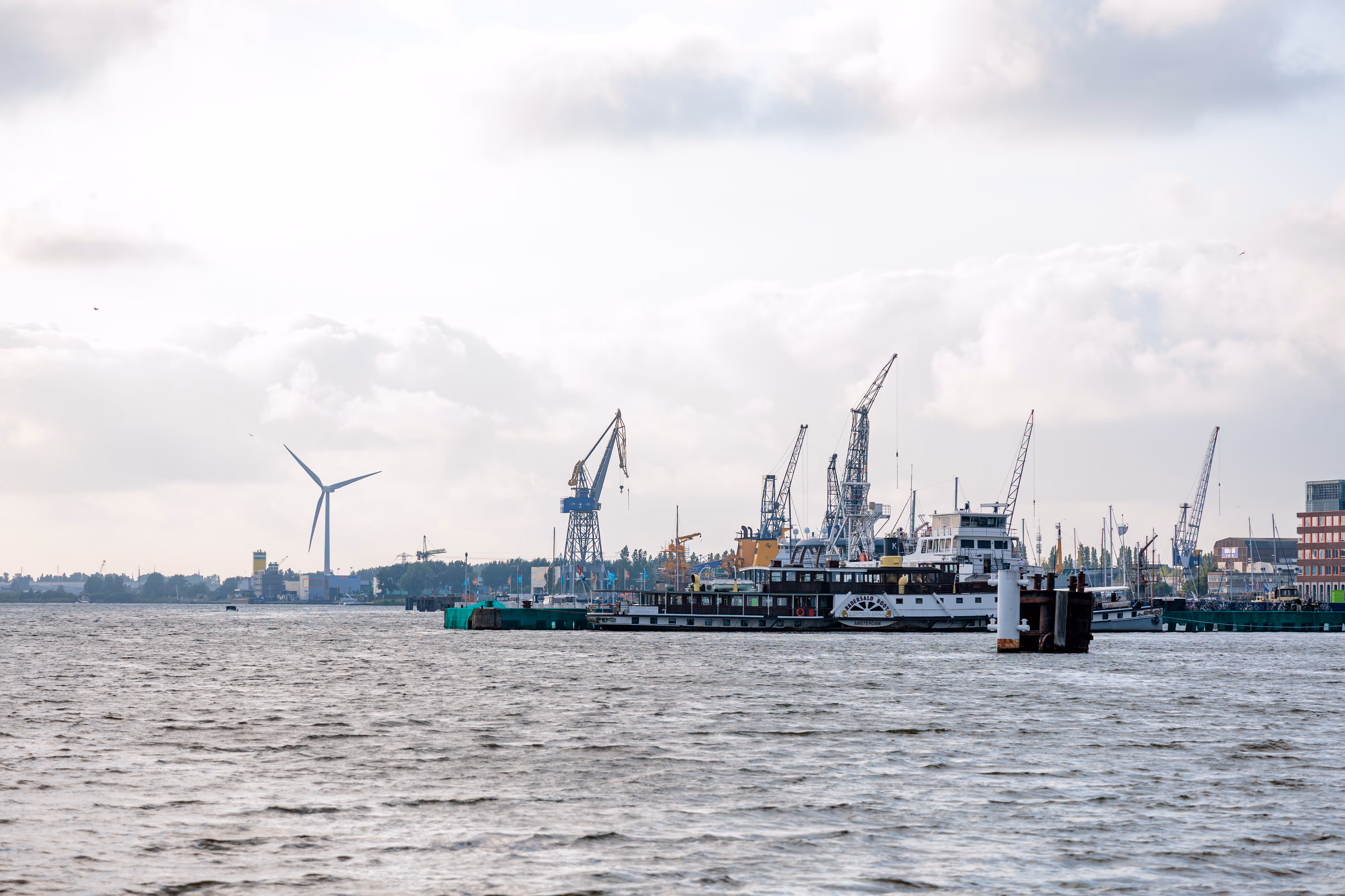 Blick über das Wasser auf den Amsterdamer Hafen mit Schiffen, Kränen und einer Windturbine unter Wolken 