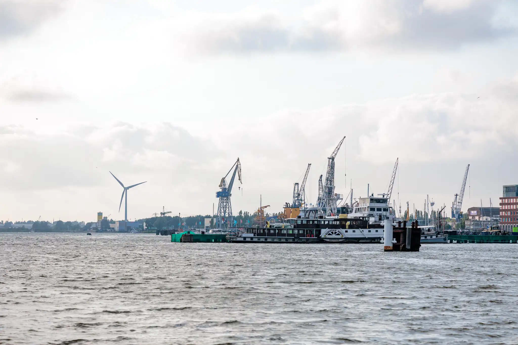 Blick über das Wasser auf den Amsterdamer Hafen mit Schiffen, Kränen und einer Windturbine unter Wolken